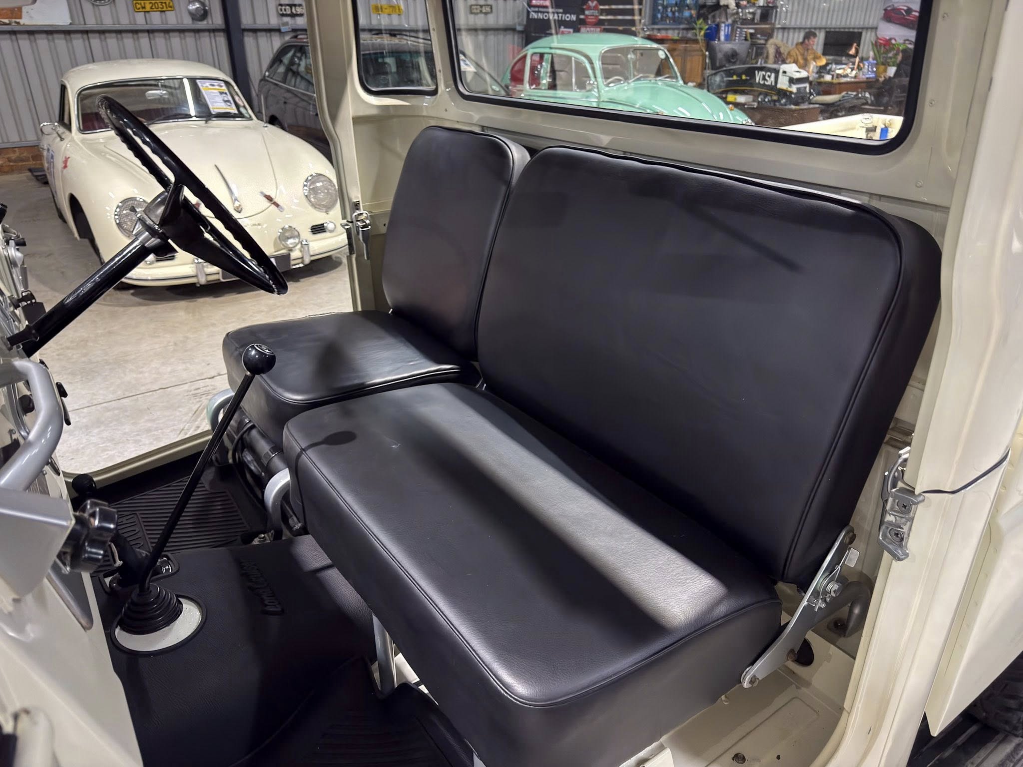Interior of a vintage vehicle with a black bench seat, steering wheel, and gear lever; showroom with classic cars visible through the window behind.