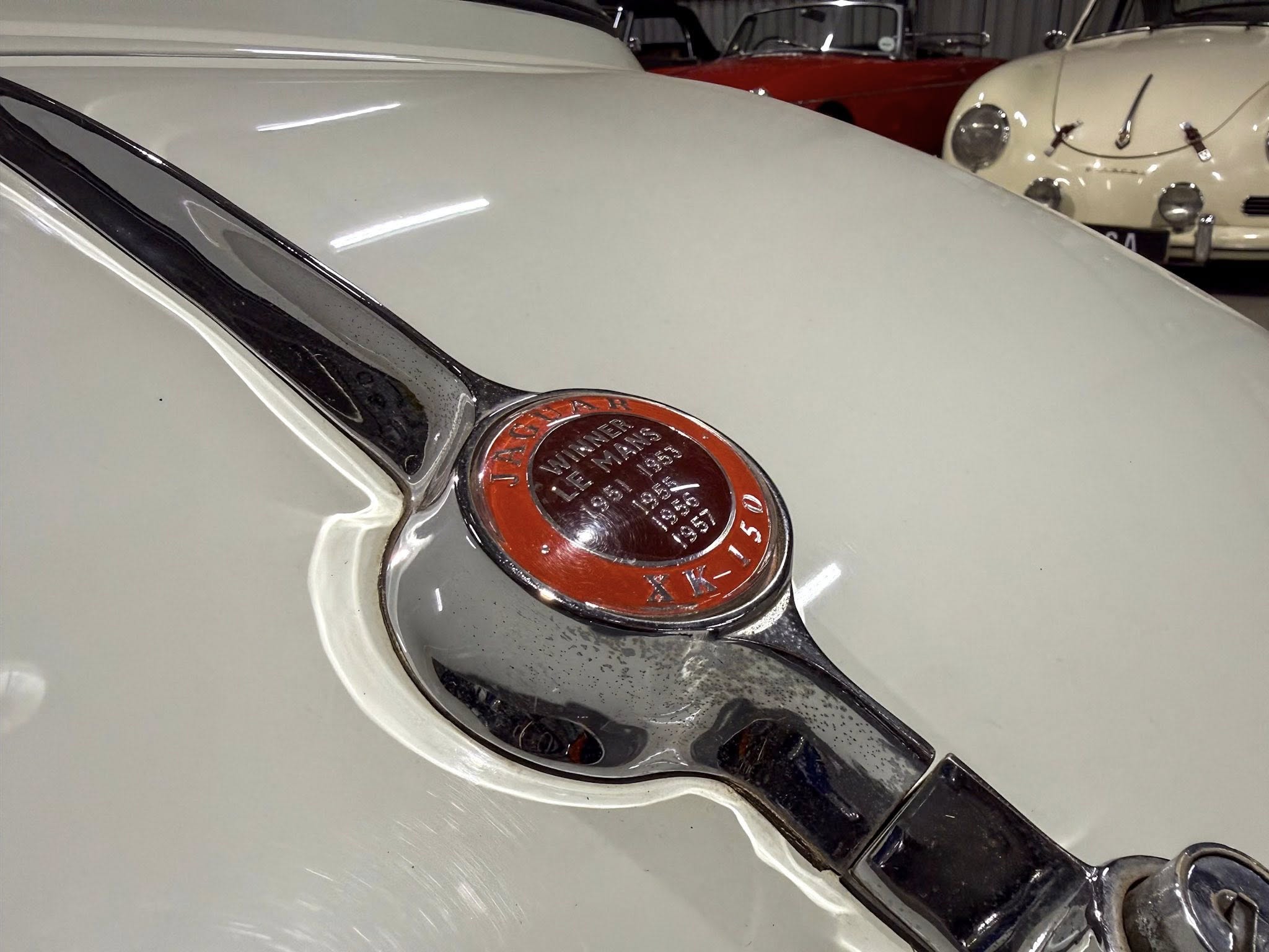 Close-up of a vintage car hood with a red circular emblem and chrome trim, with other classic cars in the background.