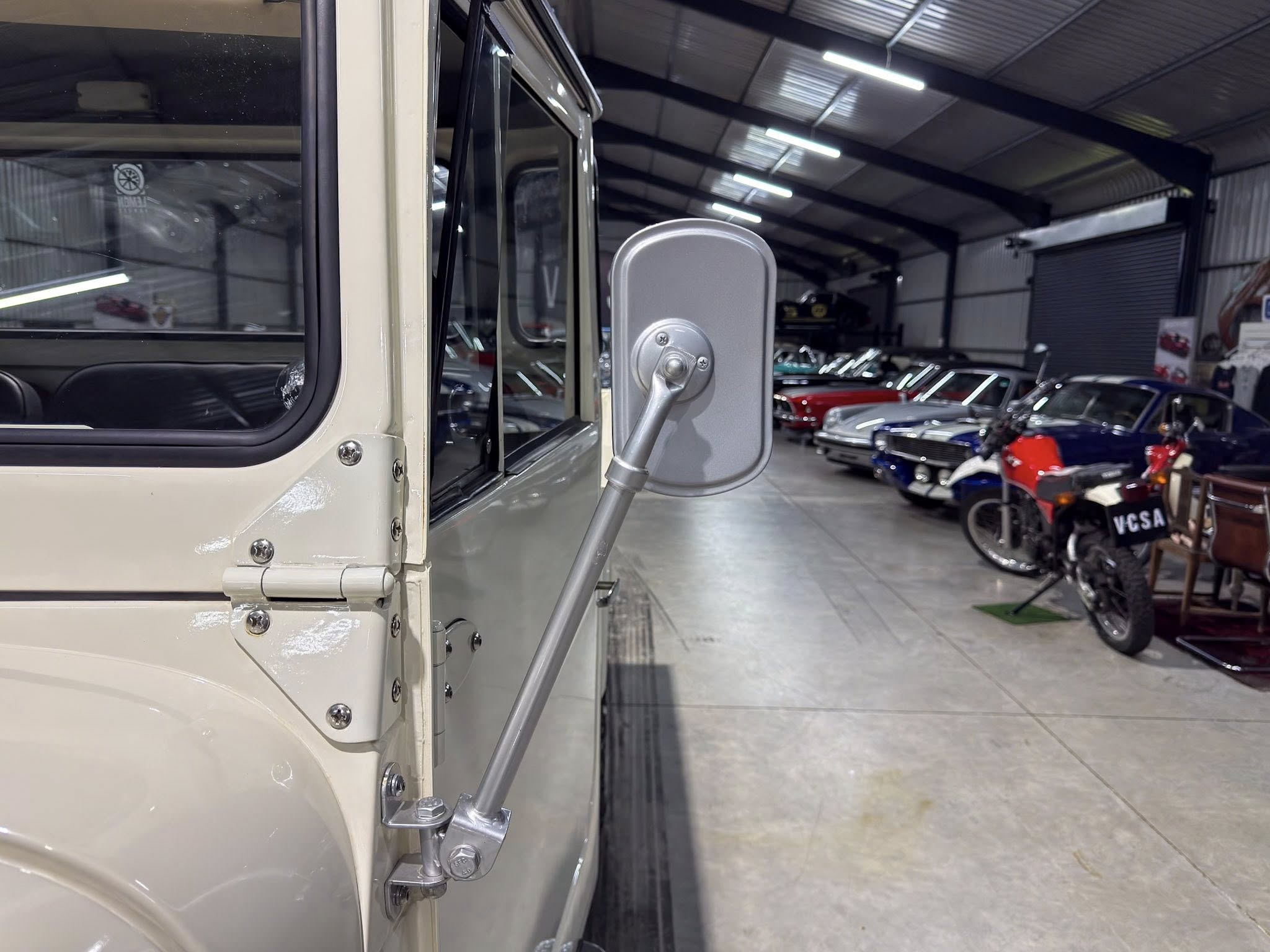 Close-up of a cream-colored vintage truck door and round side mirror in a showroom with classic cars.