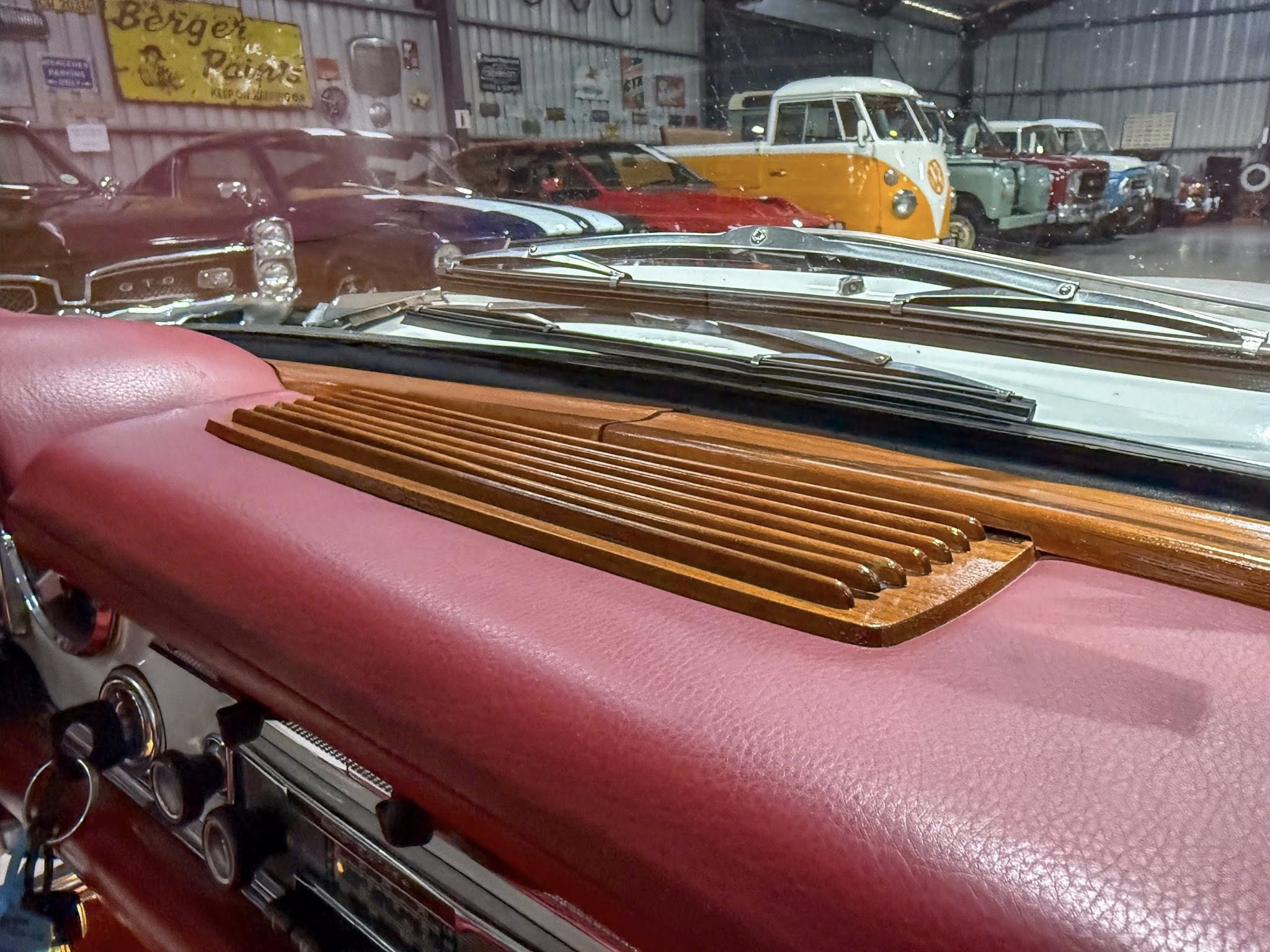 View from inside a vintage car looking out at a row of classic cars in a cluttered workshop or showroom garage.