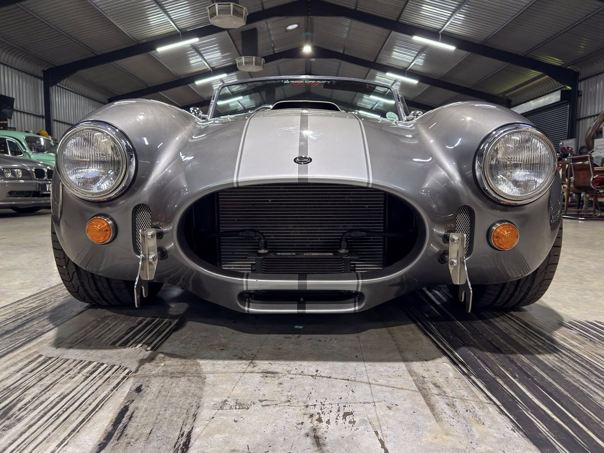 Front view of a silver vintage sports car with racing stripes in a workshop/garage.