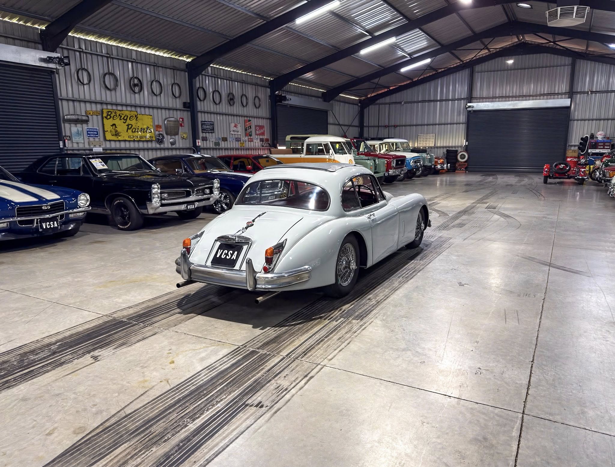 Indoor showroom of vintage cars, with a light gray classic car in the foreground and a line of colorful heritage vehicles along corrugated metal walls with retro signs on display.
