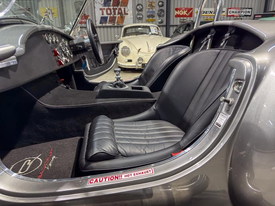 Two-seat vintage sports car interior with black leather seats, manual gear lever, and a showroom full of classic cars in the background.