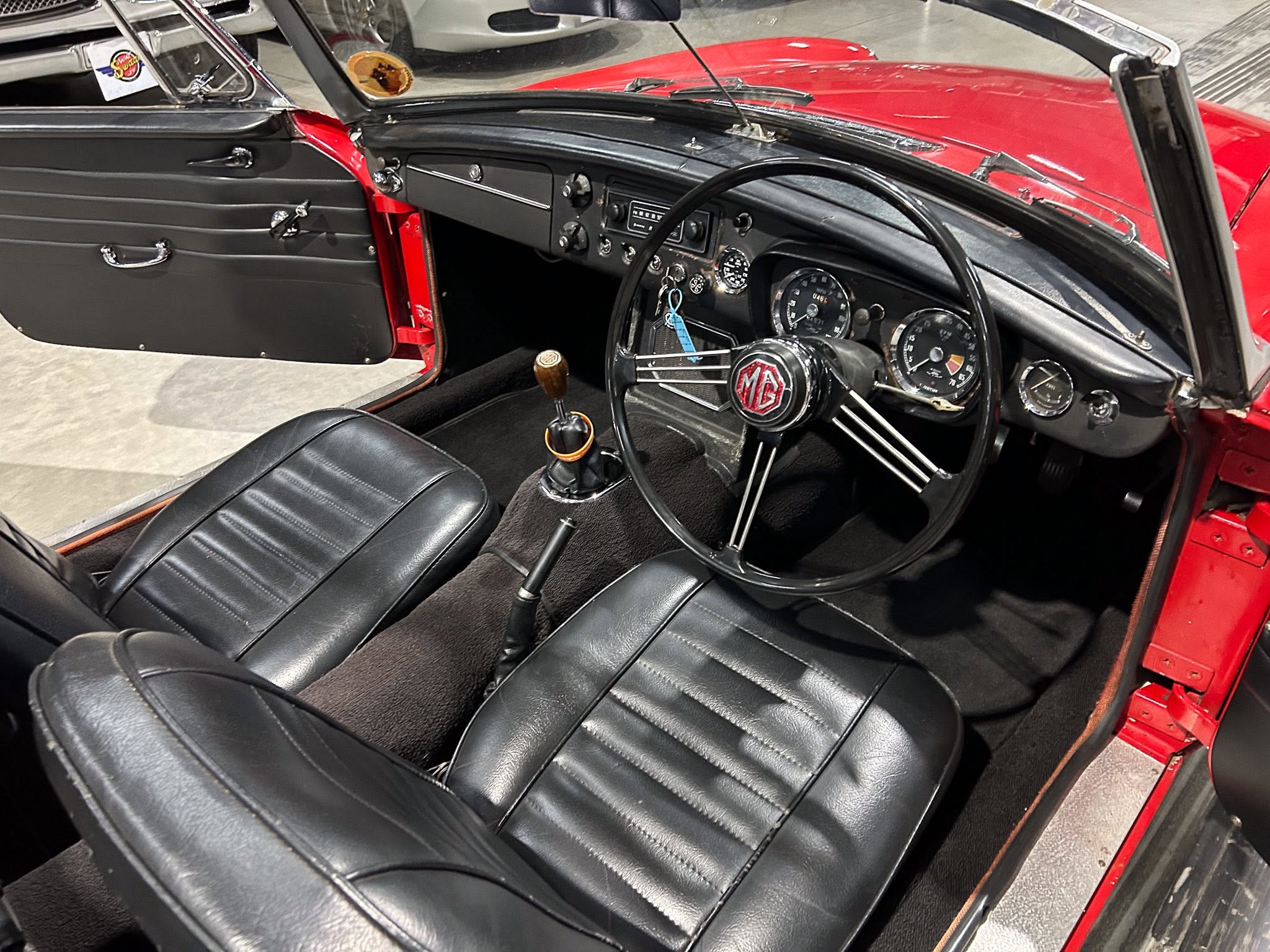 Interior of a classic red convertible: black leather seats, dashboard with round gauges, wooden gear knob, and MG-emblem steering wheel.