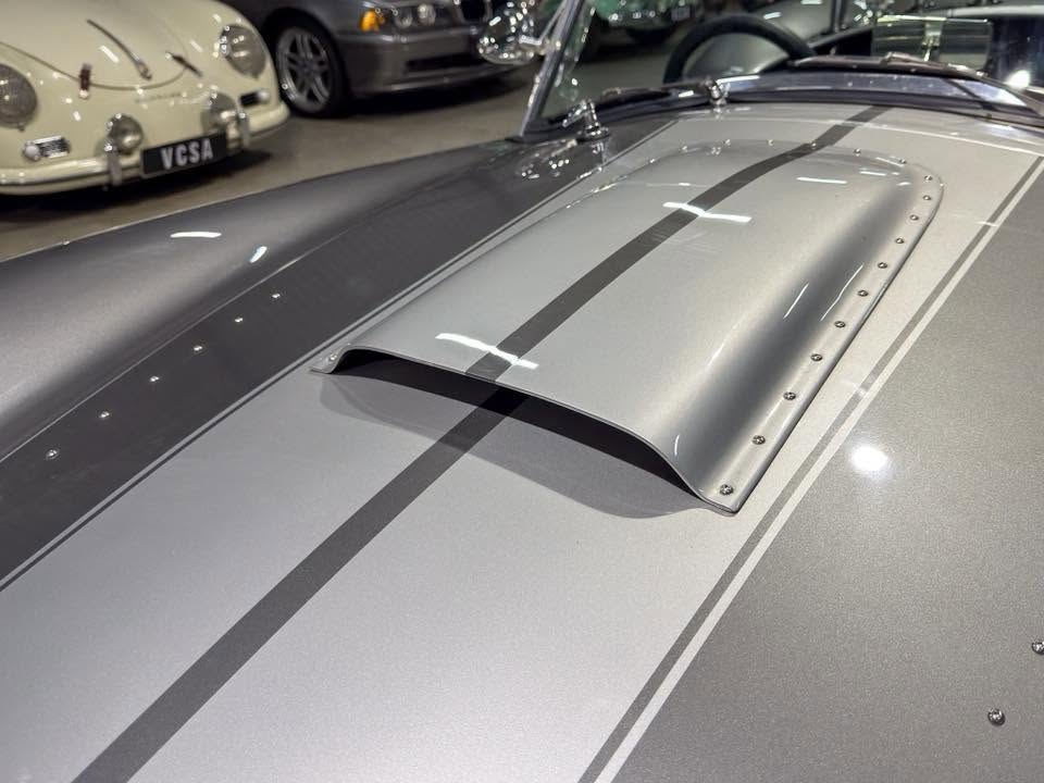 Close-up of a silver vintage car hood with a raised air scoop and racing stripes in a showroom.