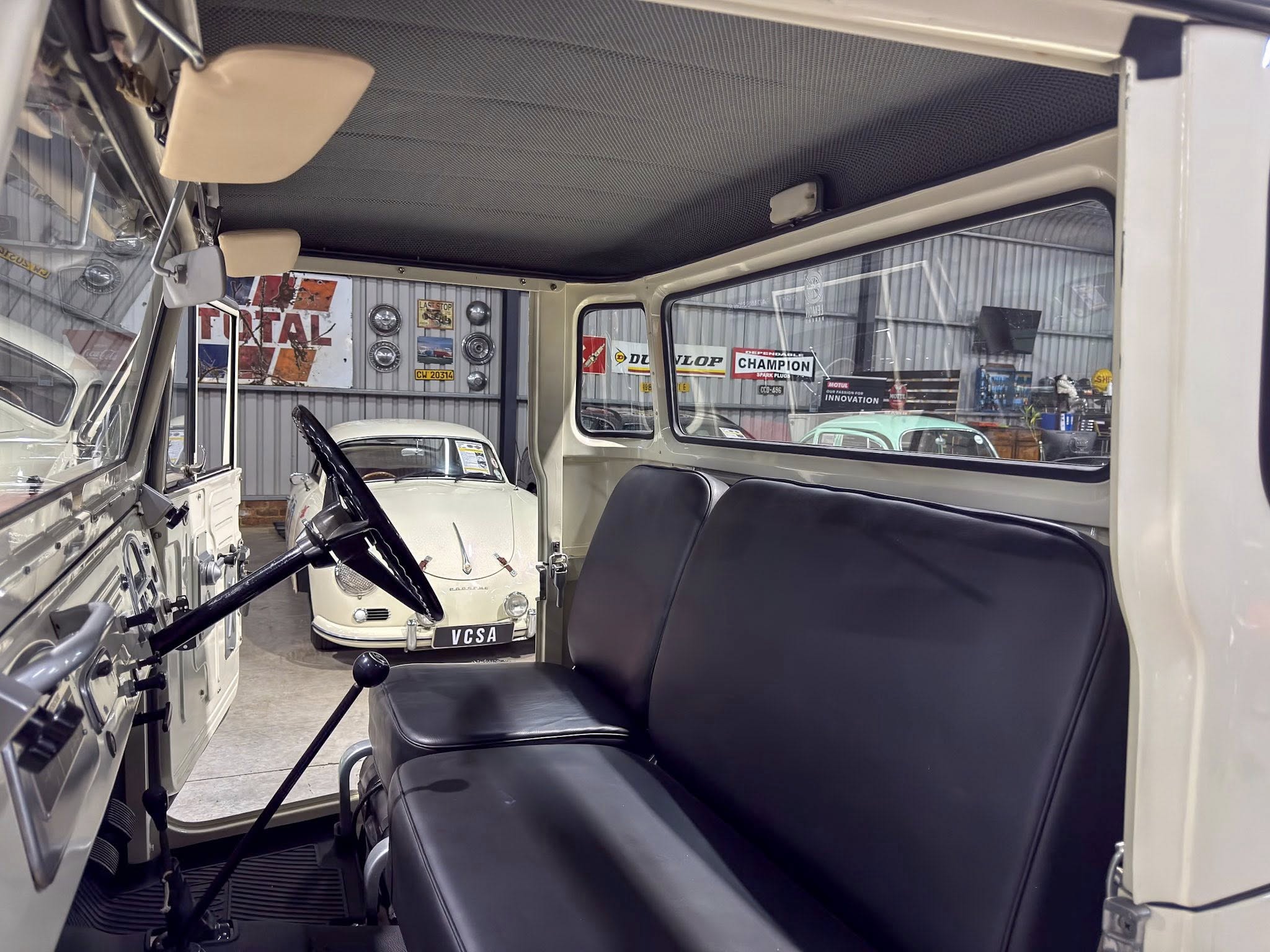 Interior view of a vintage car showing the driver’s seat and steering wheel, with a cream classic car visible through the windshield in a showroom.