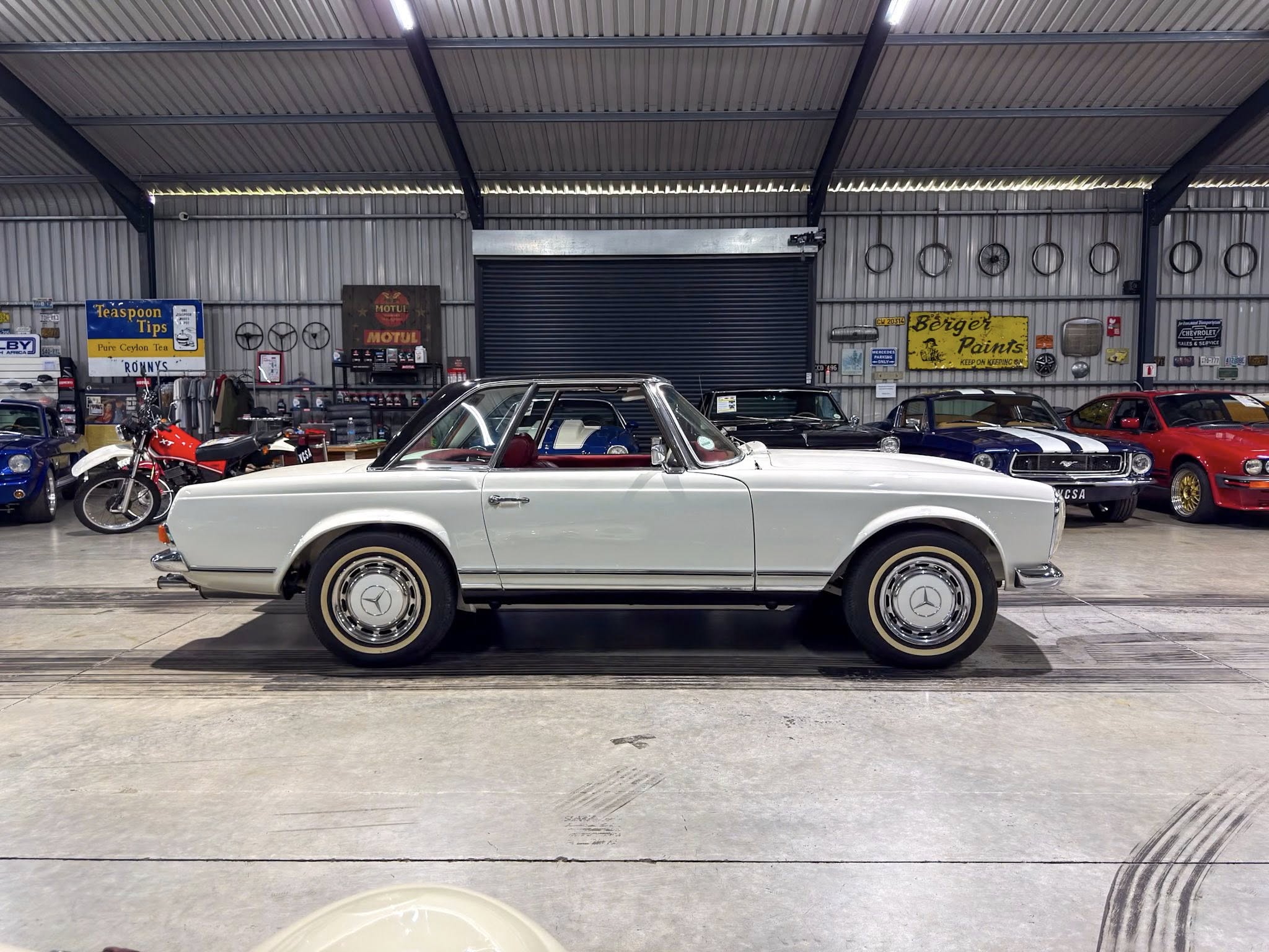 Inside a vintage car showroom with a white classic car in the foreground and other classic vehicles, signs, and tires on the walls behind.