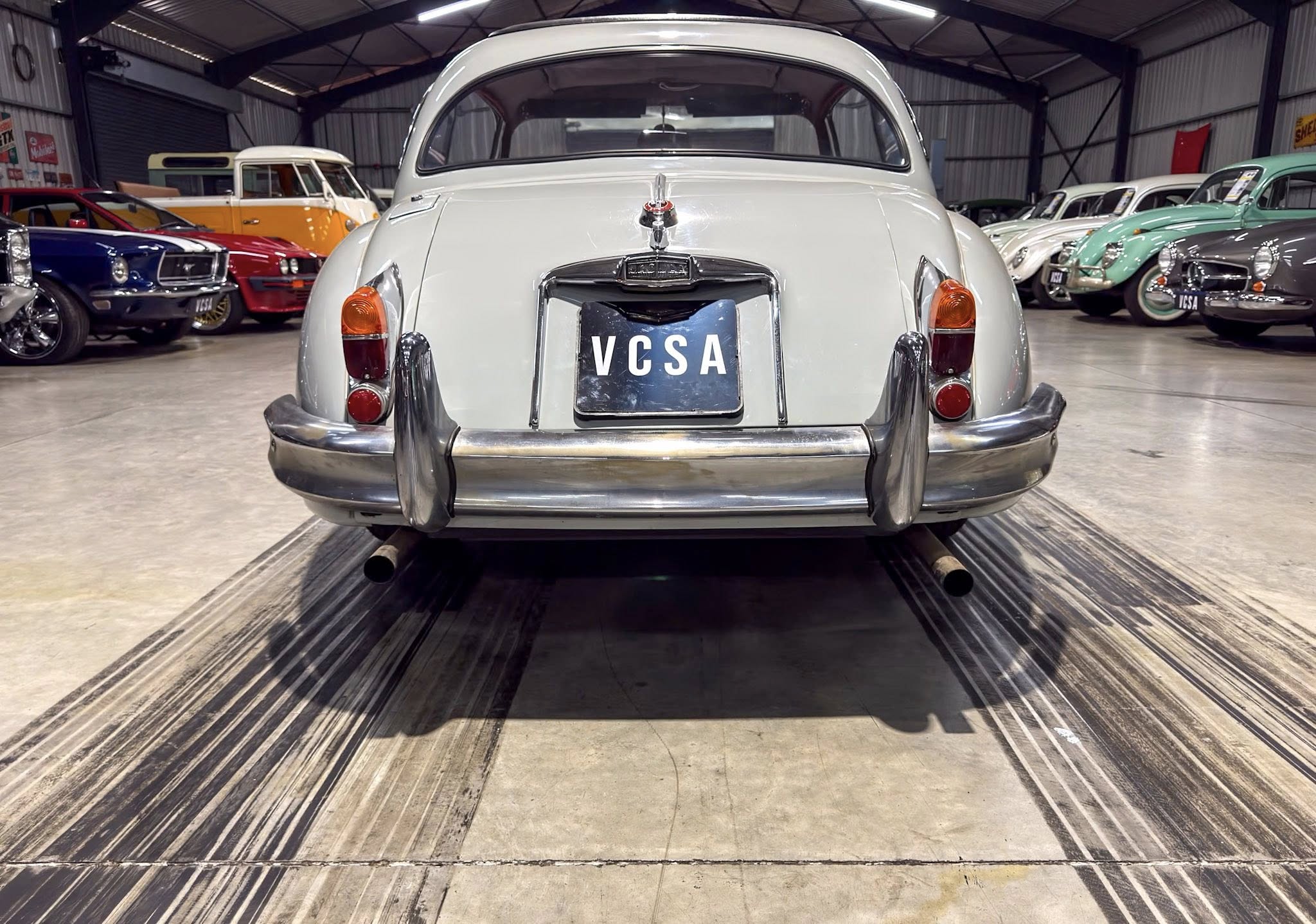 Rear view of a light gray vintage car on a showroom floor, surrounded by colorful classic cars in a well-lit warehouse.