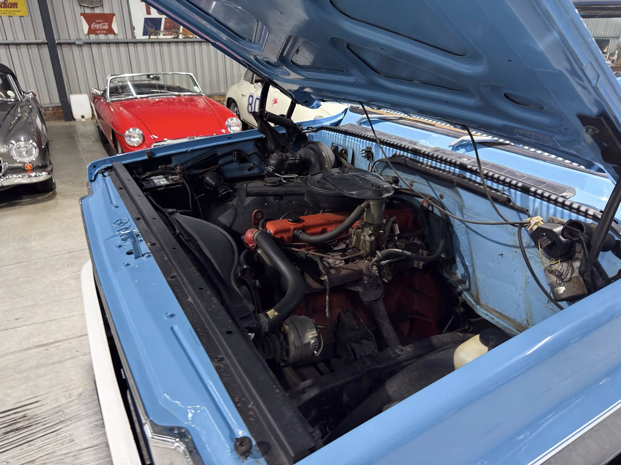 Blue vintage car with its hood raised, exposing the engine in a showroom with a red convertible in the background.