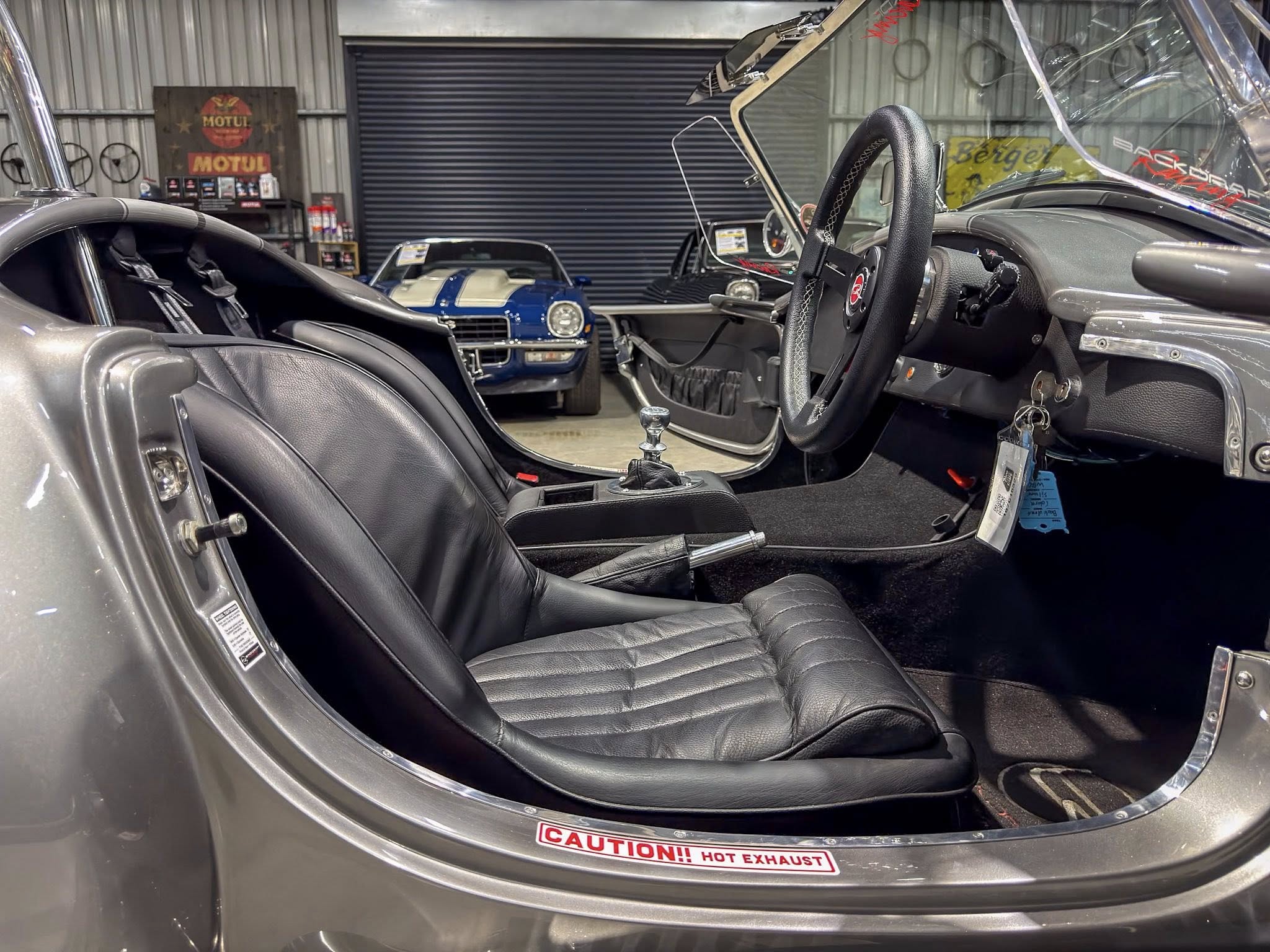 Classic car interior with black leather seats, steering wheel, and gear shifter; a blue vintage car is visible in the background inside a garage showroom.