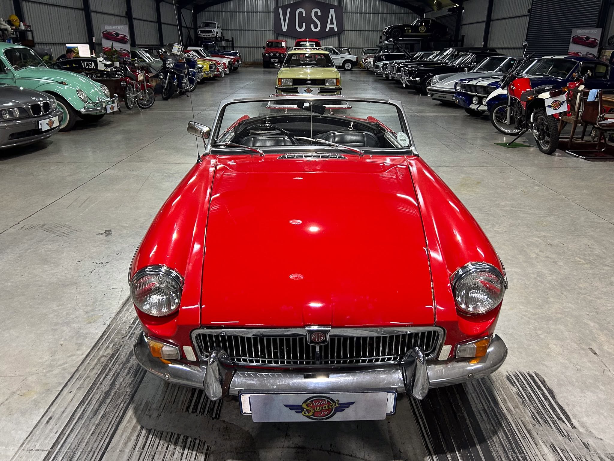 Red classic convertible viewed from the front center, inside a large showroom filled with vintage cars and motorcycles on both sides.