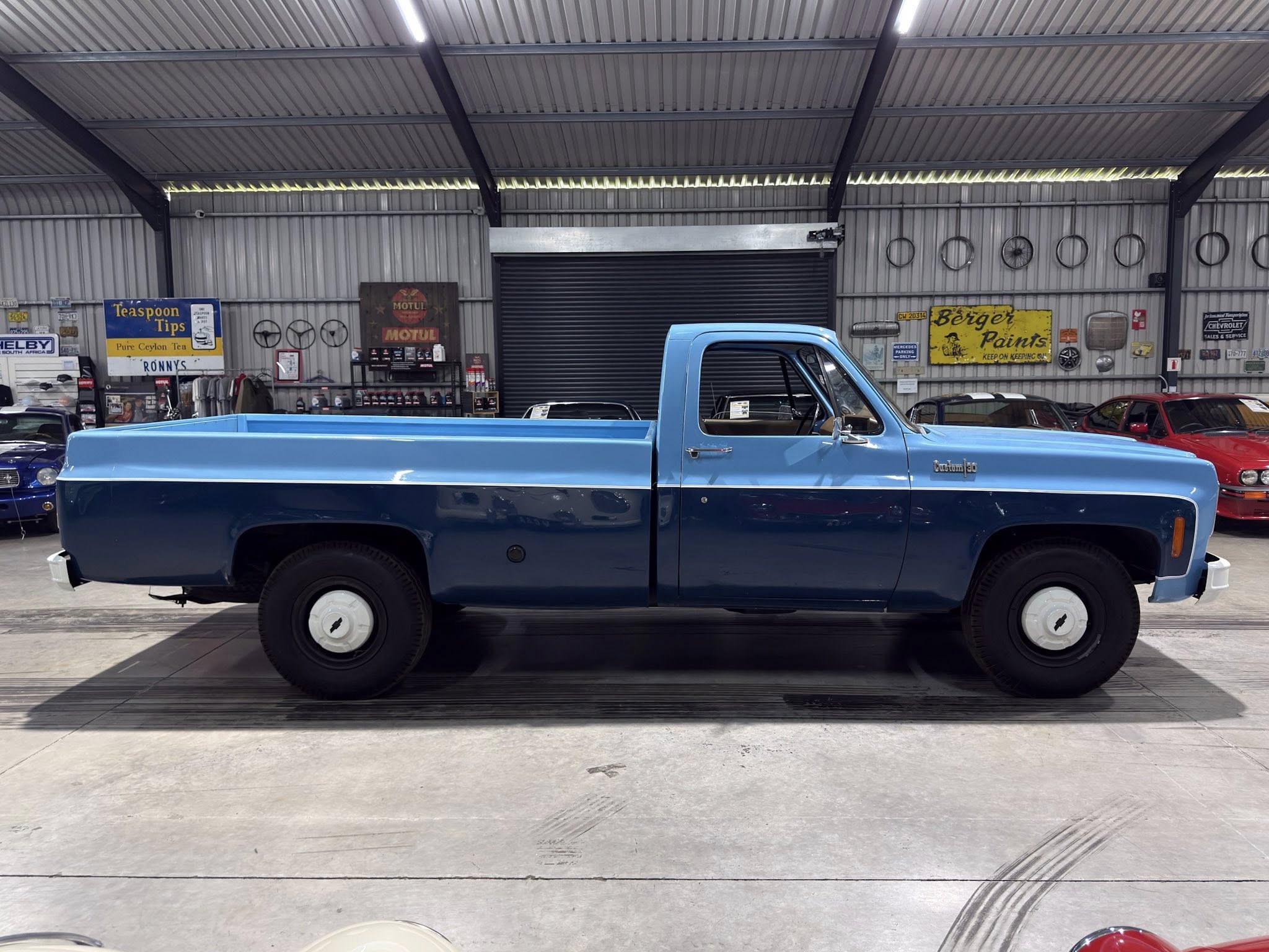 Blue classic pickup truck photographed inside a metal‑walled workshop with vintage signs on the back wall.