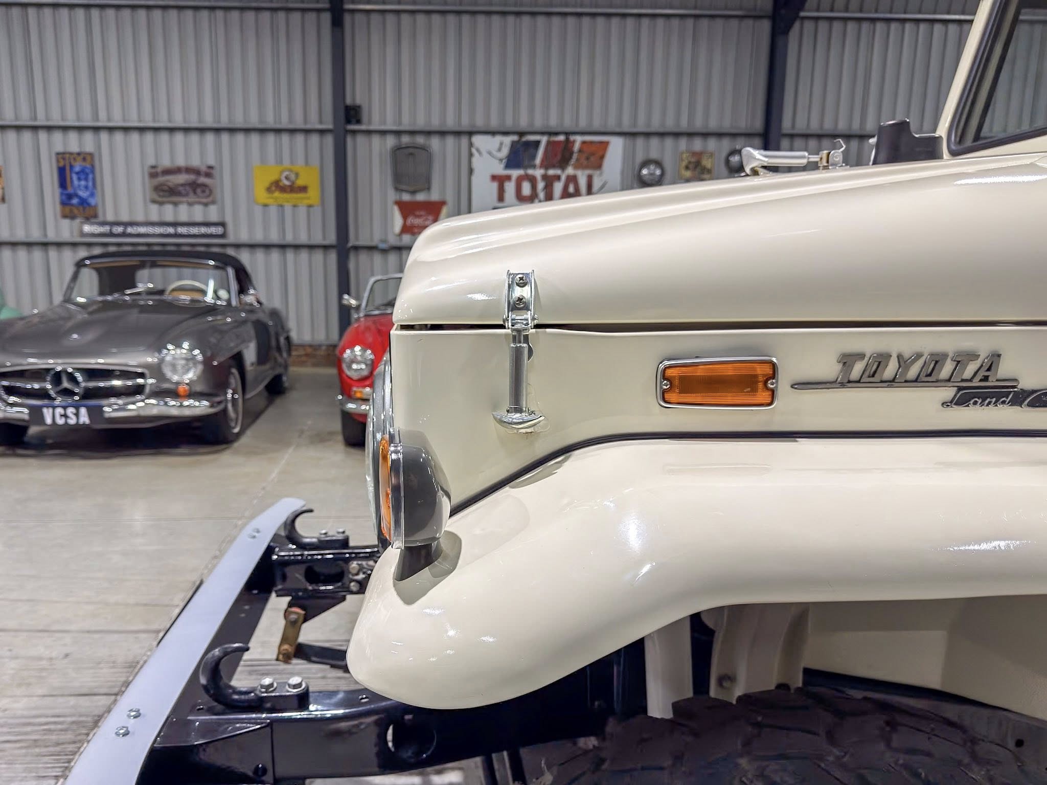 Close-up of a cream vintage car's front fender and headlight in a showroom with other classic cars in the background.