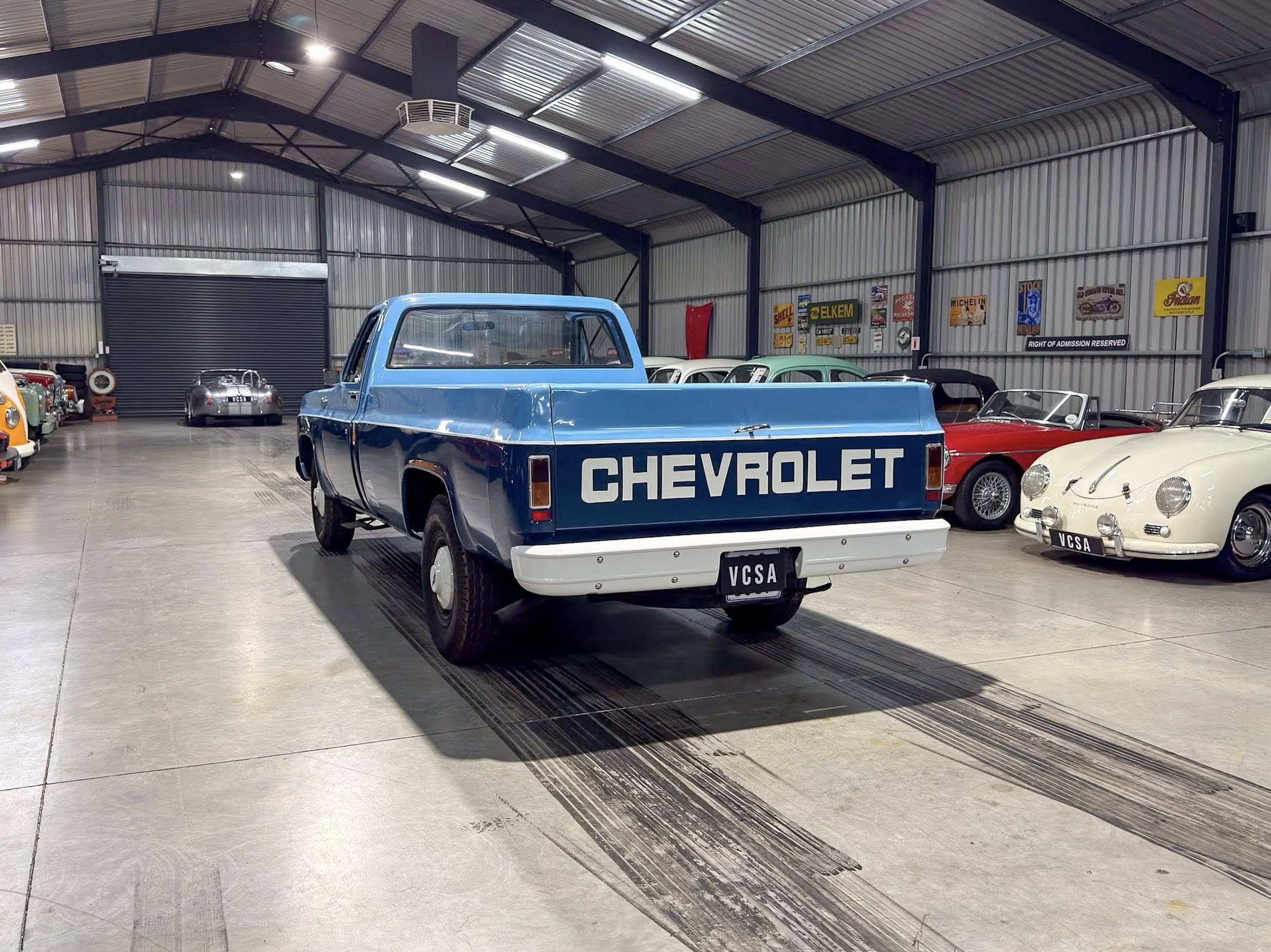 Blue Chevrolet pickup parked inside a vintage car showroom with other classic cars along the right wall and metal industrial interior behind.