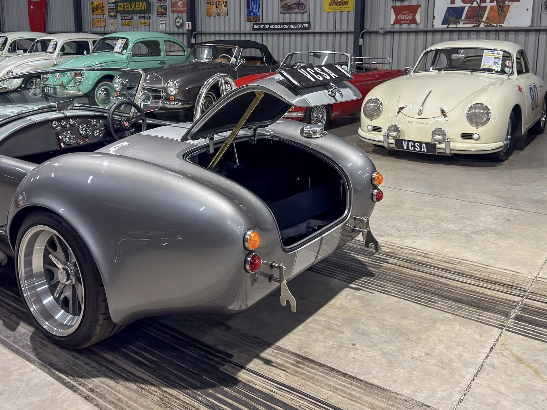 Classic silver sports car with rear hatch open in a vintage car showroom, other retro cars visible in background against a garage interior.