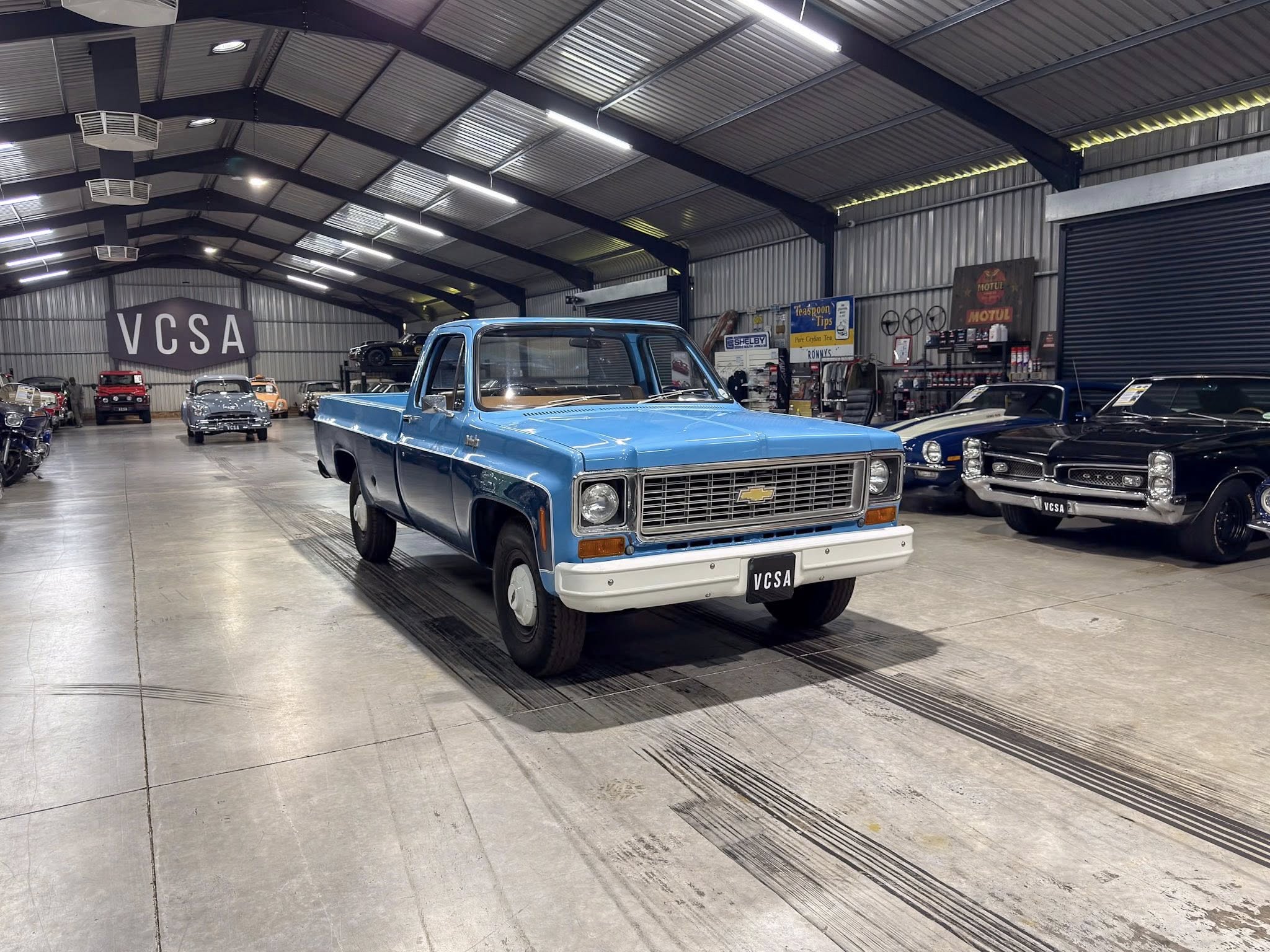 Blue vintage pickup truck in a classic car showroom with other retro cars and a 'VCSA' sign in the background.