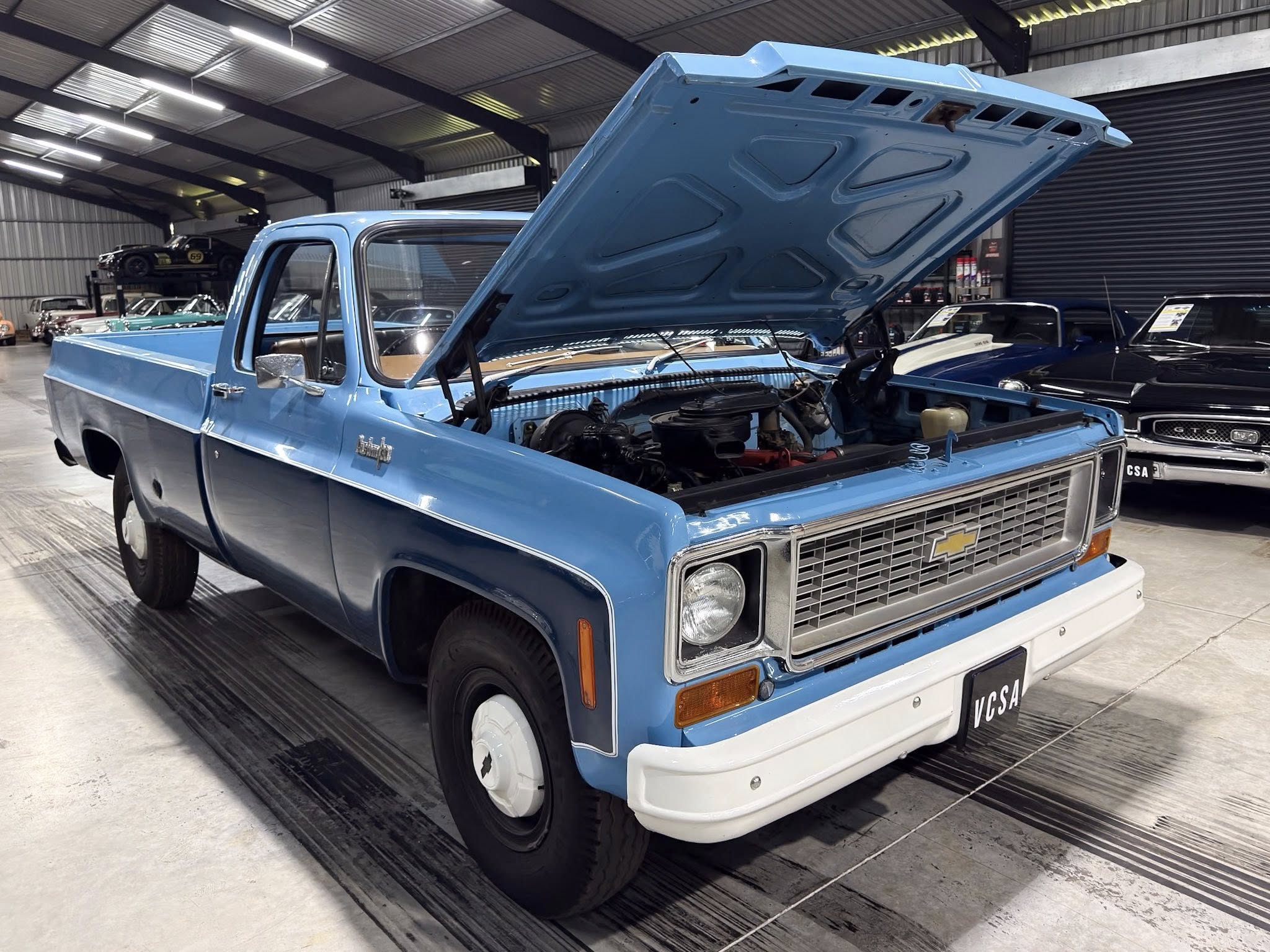 Blue vintage Chevrolet pickup with its hood open in a showroom, other classic cars visible in the background