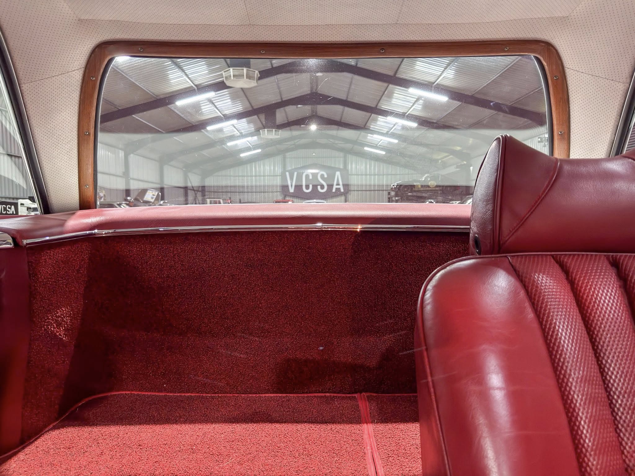 Interior of a vintage car with red leather seats and wood-trimmed rear window, view through the window shows a warehouse with 'VCSA' sign.