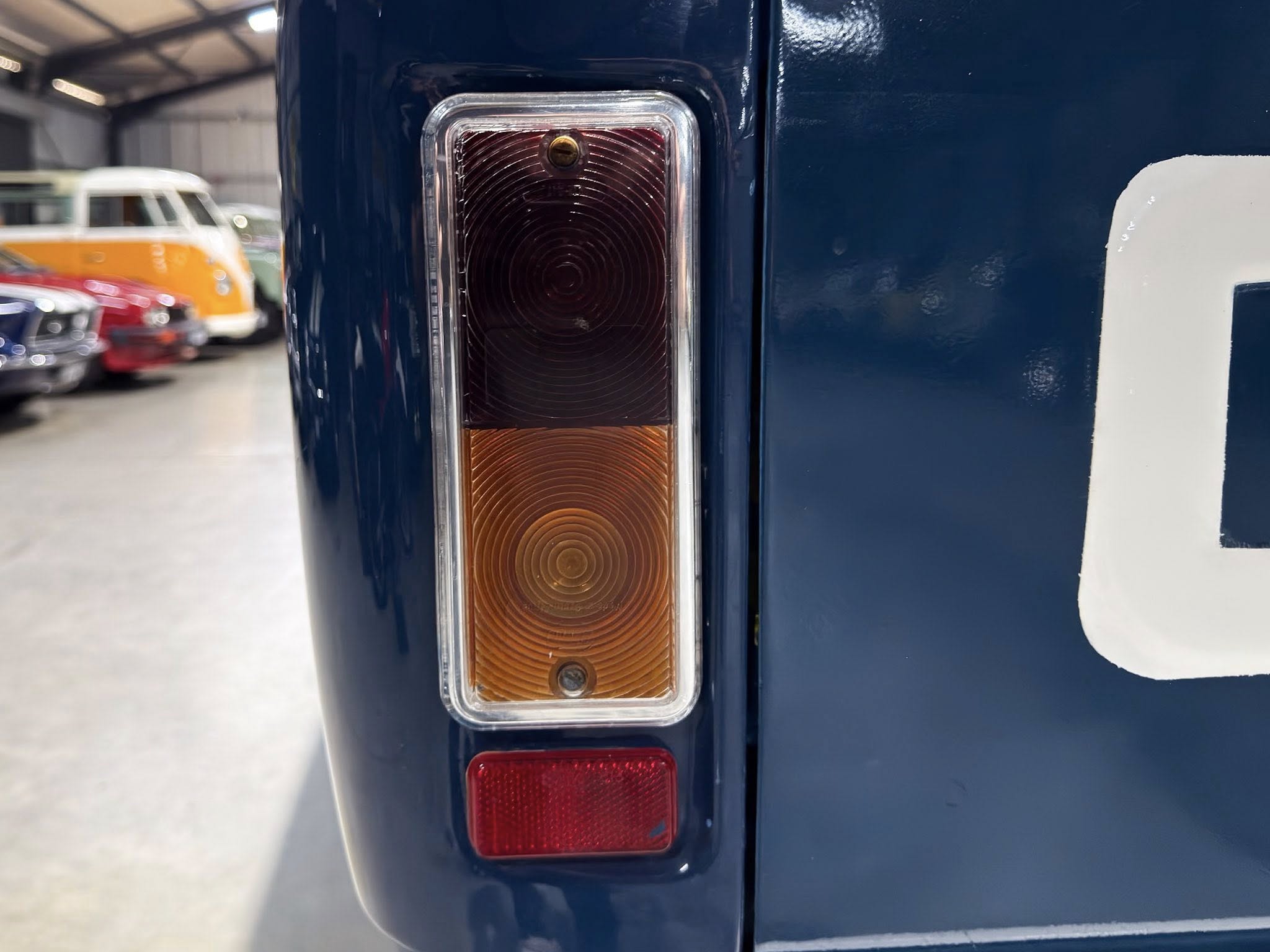 Close-up of a blue vehicle's rectangular tail light with orange indicator lenses in a workshop/garage. In the background, other cars are blurred.