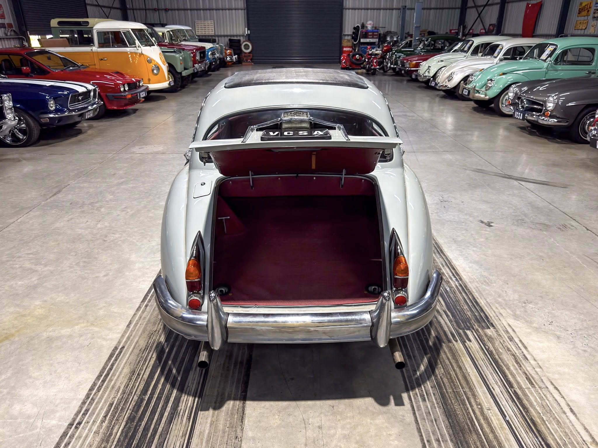 Indoor showroom filled with vintage cars; foreground shows a pale blue car with open trunk, surrounded by colorful classics on both sides.