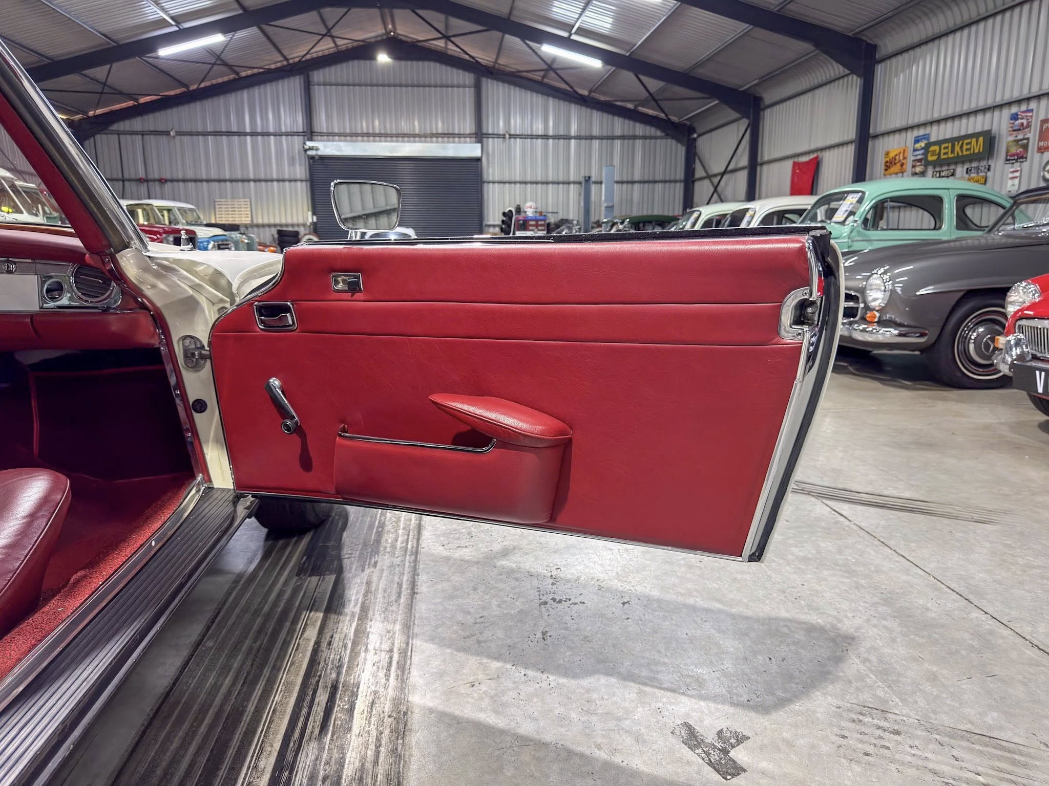 Open red car door of a vintage convertible in a showroom with other classic cars in the background.