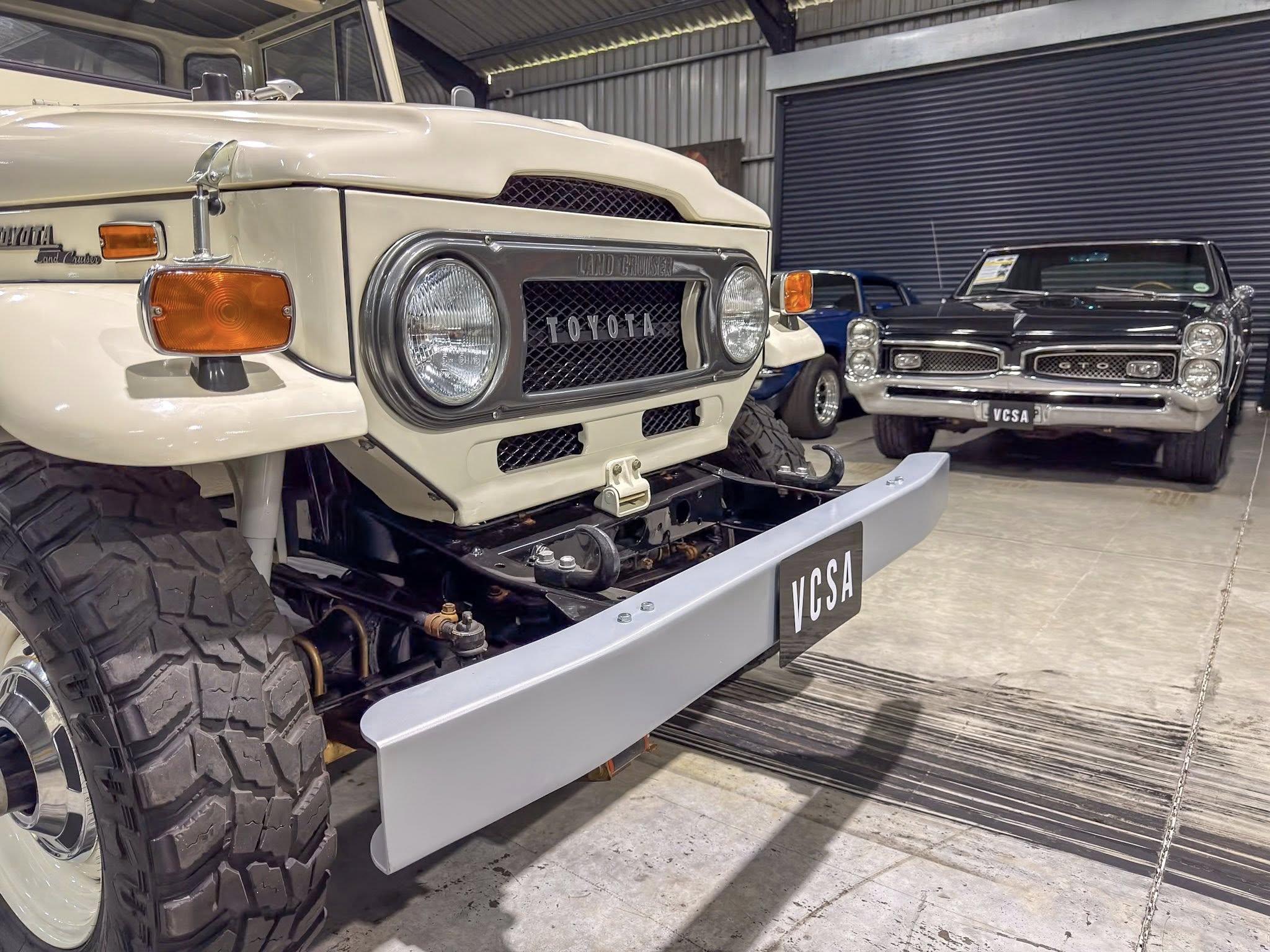 Front view of a beige Toyota Land Cruiser with oversized off-road tires on a showroom display, other classic cars visible in the background.