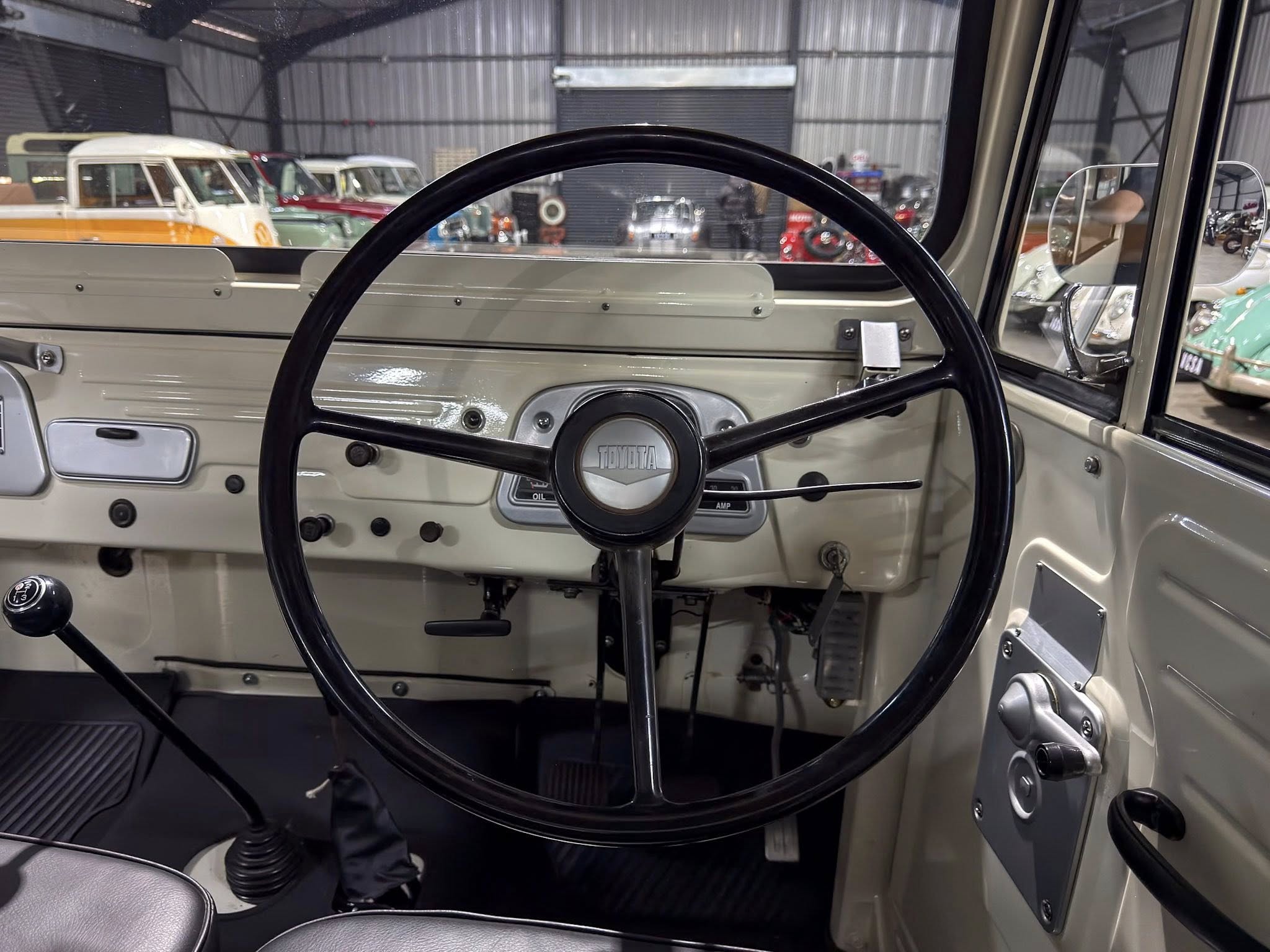 Interior of a vintage Toyota car: large black steering wheel, beige dashboard, and manual gear lever, with classic cars visible in a museum through the windshield.