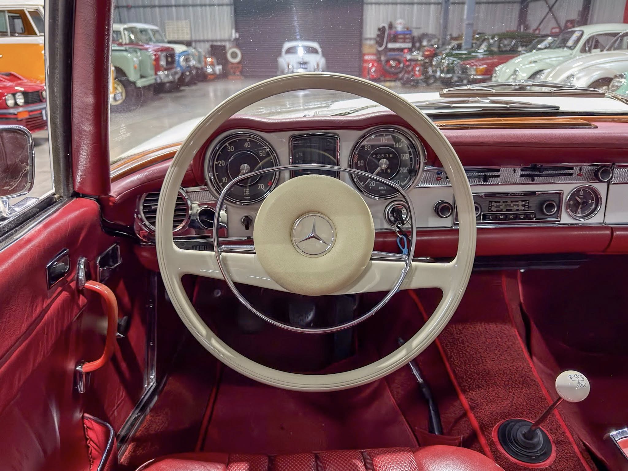 Inside view of a red vintage car’s dashboard with a cream steering wheel and classic gauges, showroom cars visible through the windshield.