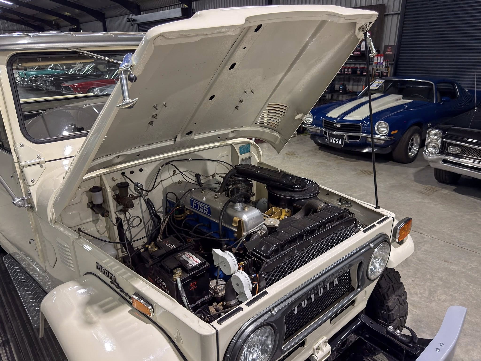 Jeep with its hood raised showing the engine, inside a showroom or workshop with classic cars in the background.