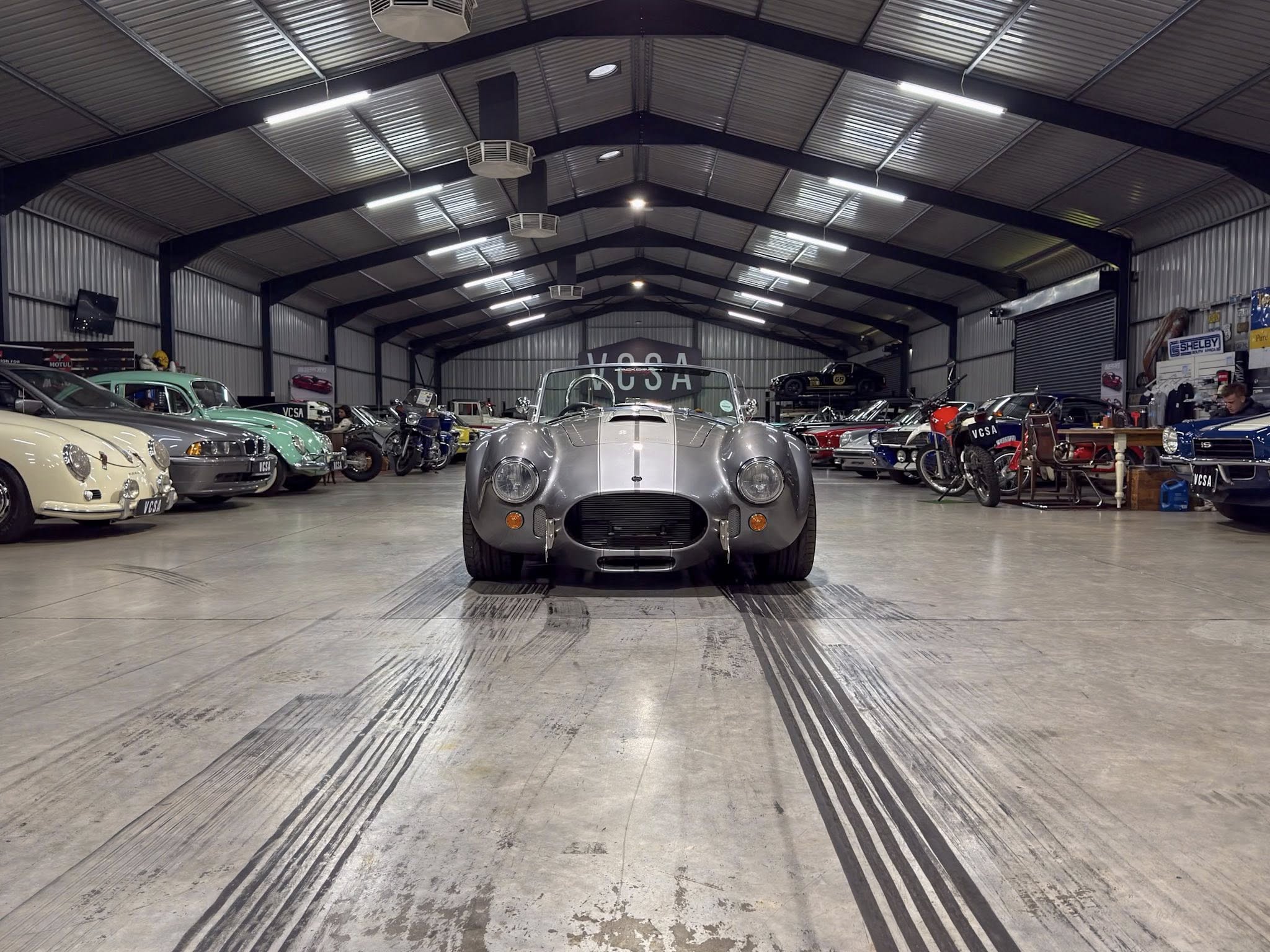 Silver vintage sports car centered in a spacious showroom, with rows of classic cars on either side.
