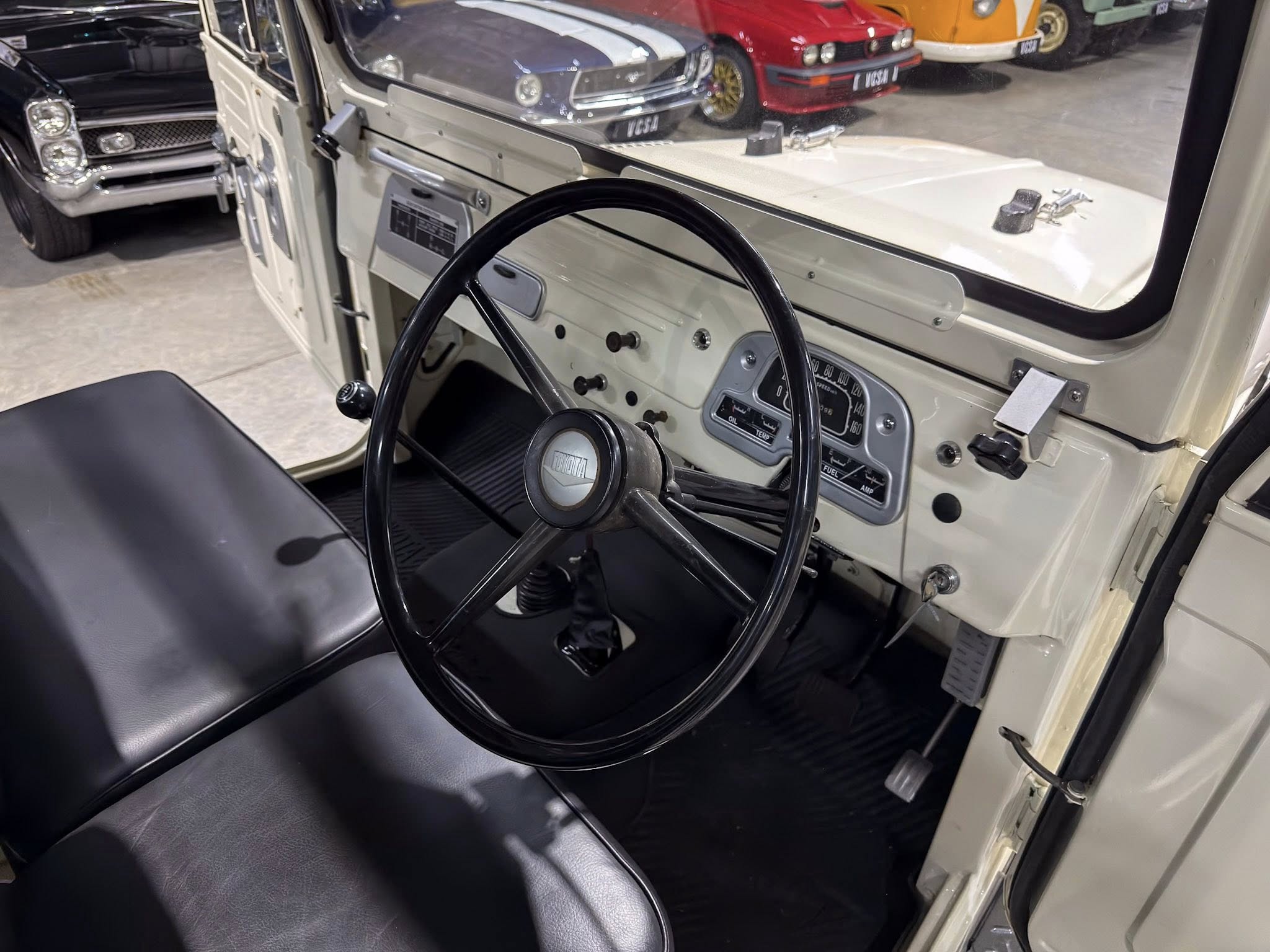 Interior of a vintage car: beige dashboard, black steering wheel, and classic gauges visible from the passenger seat, showroom with other classic cars in the background