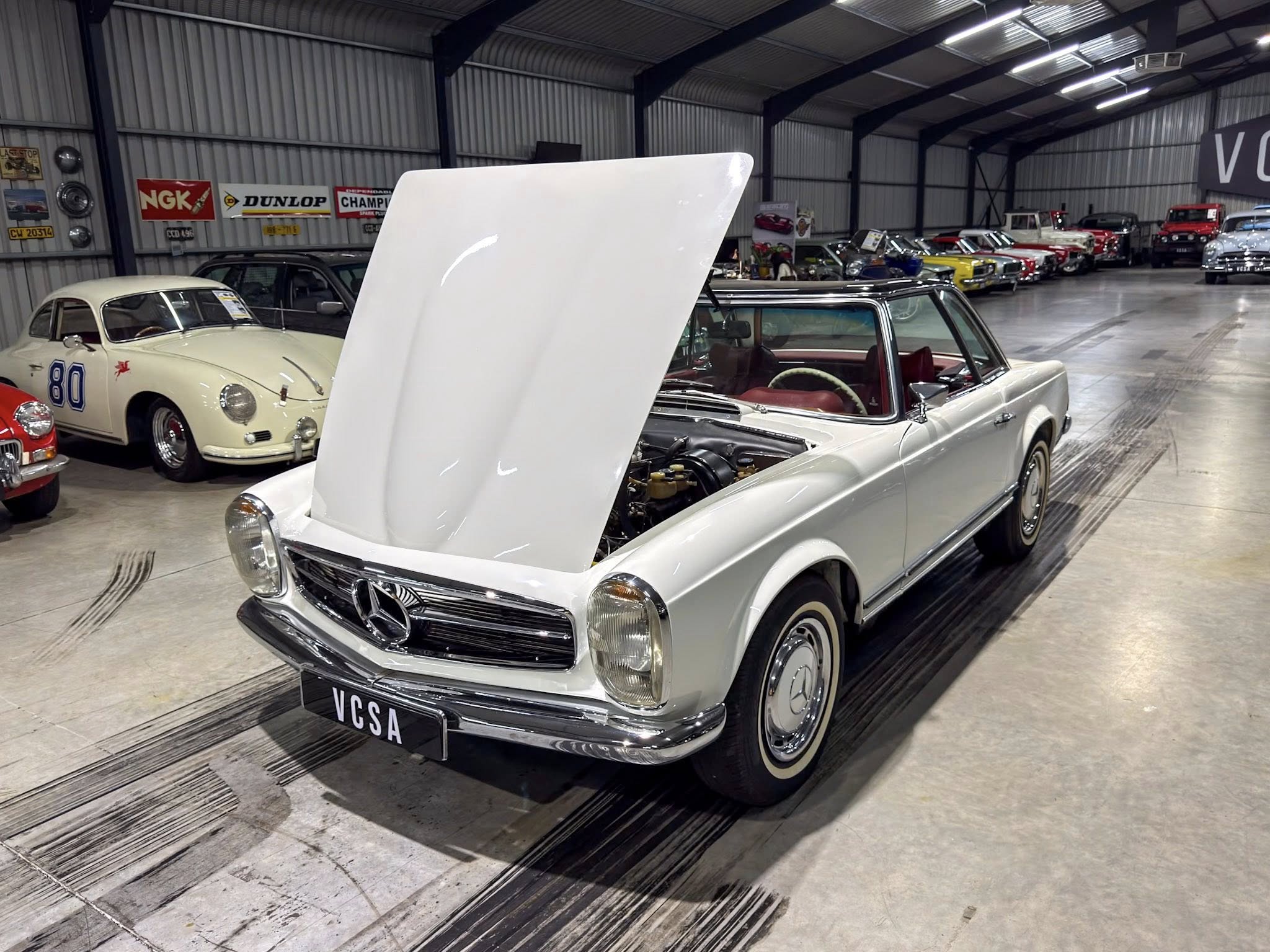 White vintage Mercedes with raised hood in a classic car showroom, rows of colorful antique cars in the background.