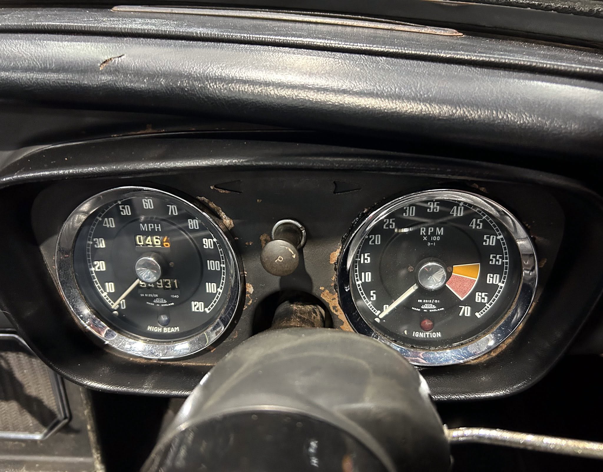 Two round analog gauges on a vintage car dashboard (speedometer on the left and tachometer on the right) with a worn black surface and visible rust spots. The left gauge shows mph and an odometer, while the right gauge shows RPM.