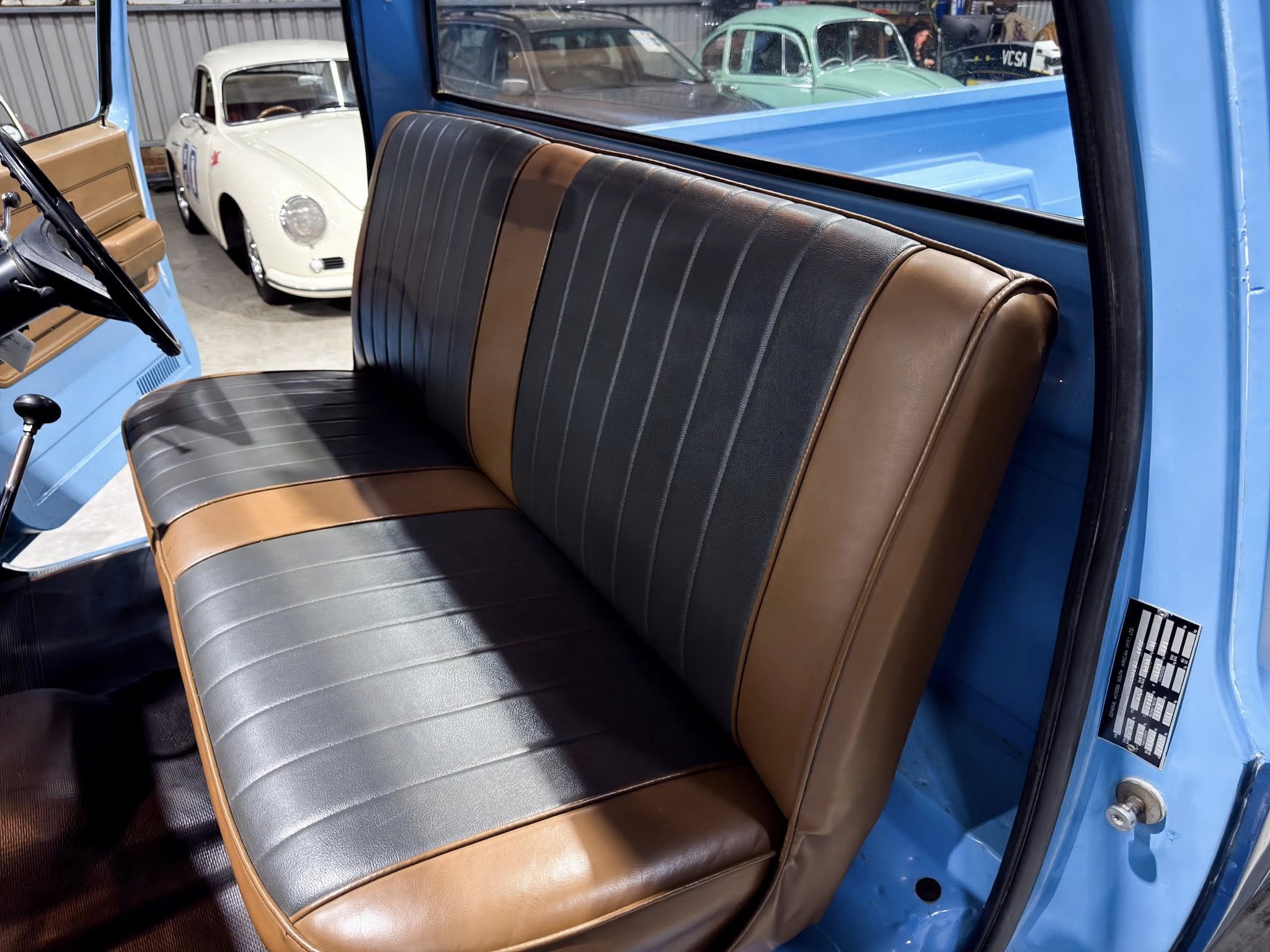 Interior of a vintage blue car with brown and black vinyl bench seat, door open, showing classic cars in a showroom behind it.