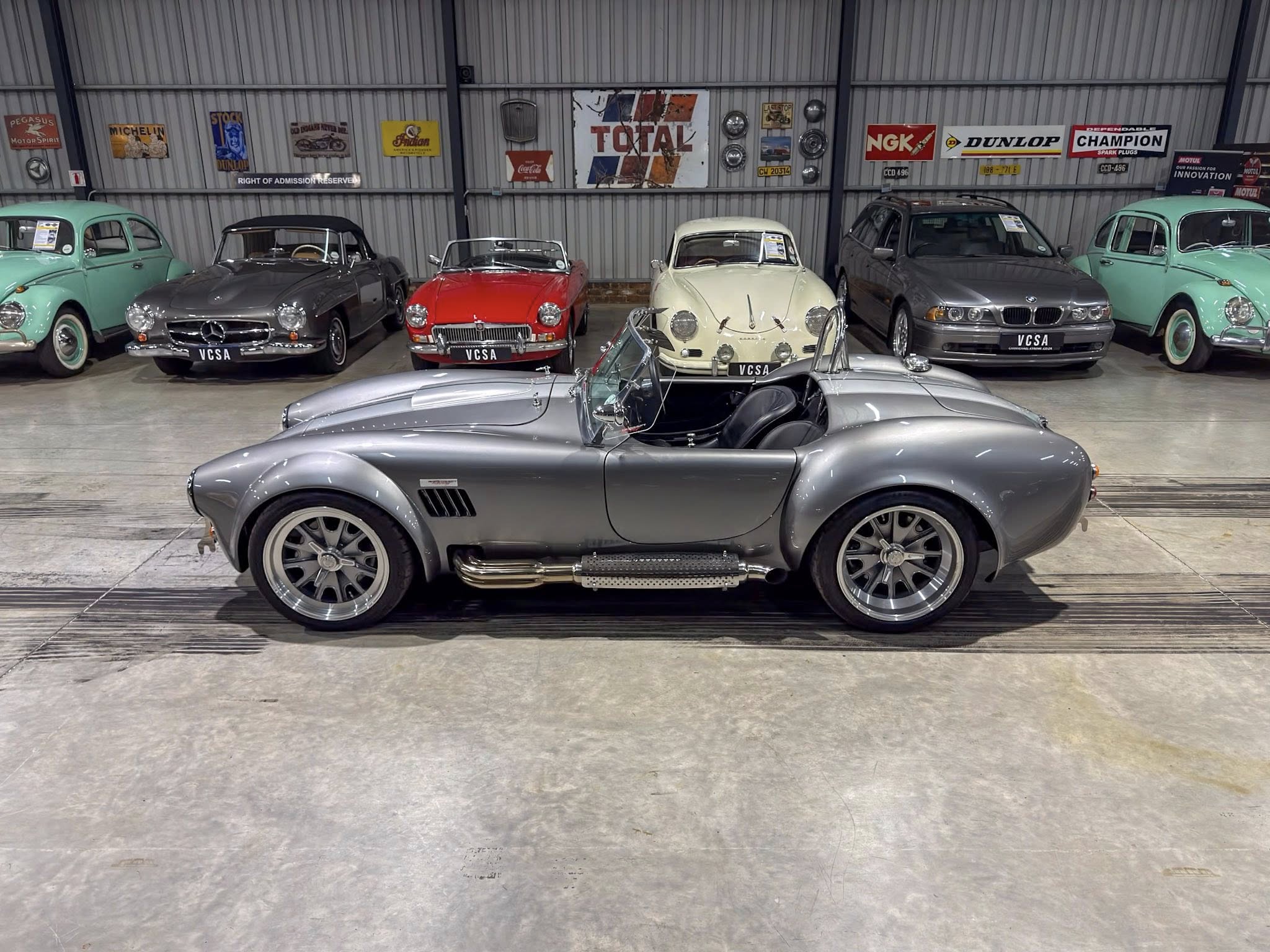 Indoor showroom with classic cars lined up; foreground silver convertible, other vintage cars in pastel and gray behind.