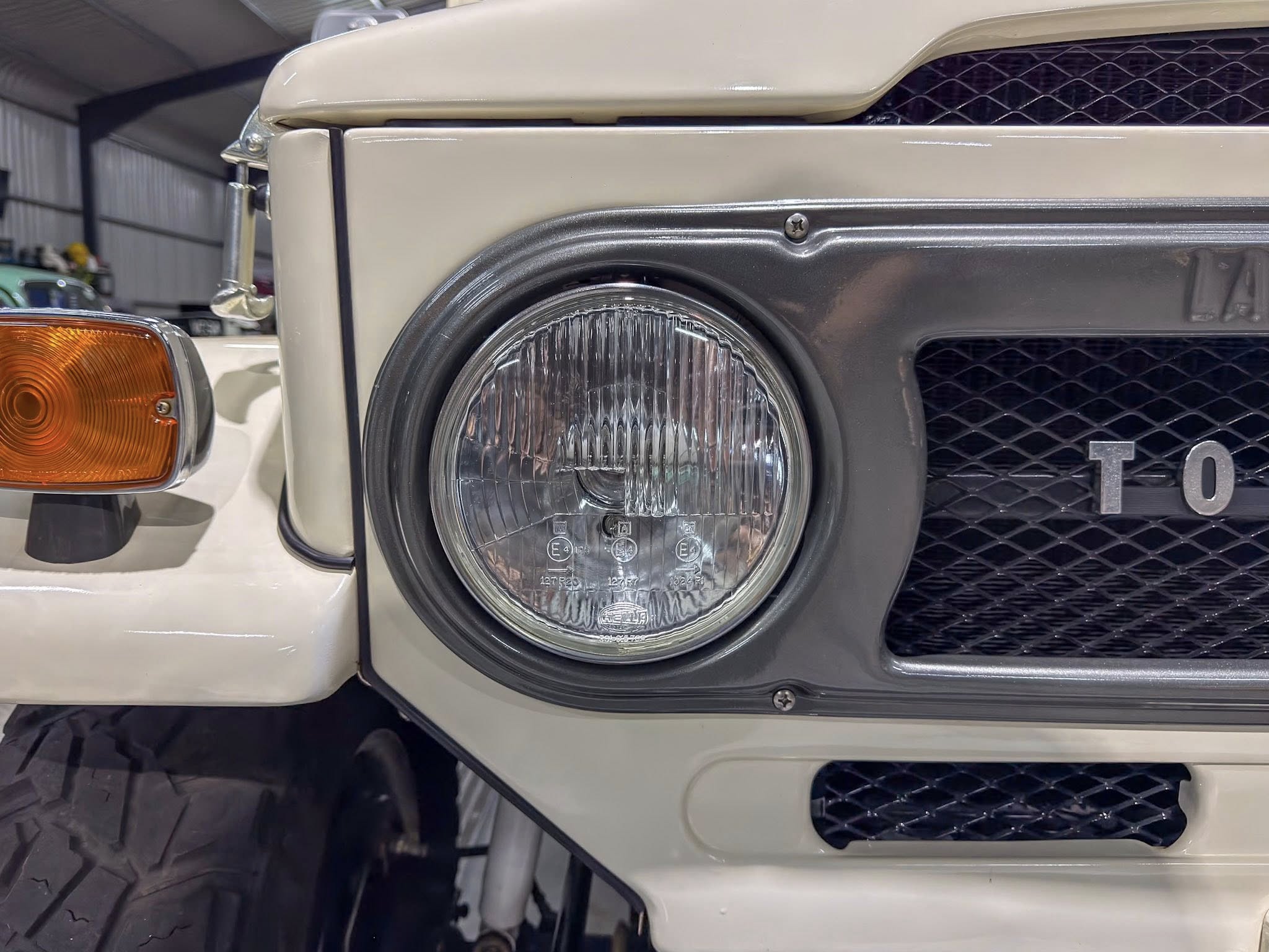 Close-up of the front left corner of a beige vintage car, showing the round headlight, orange indicator lamp, and chrome bumper.