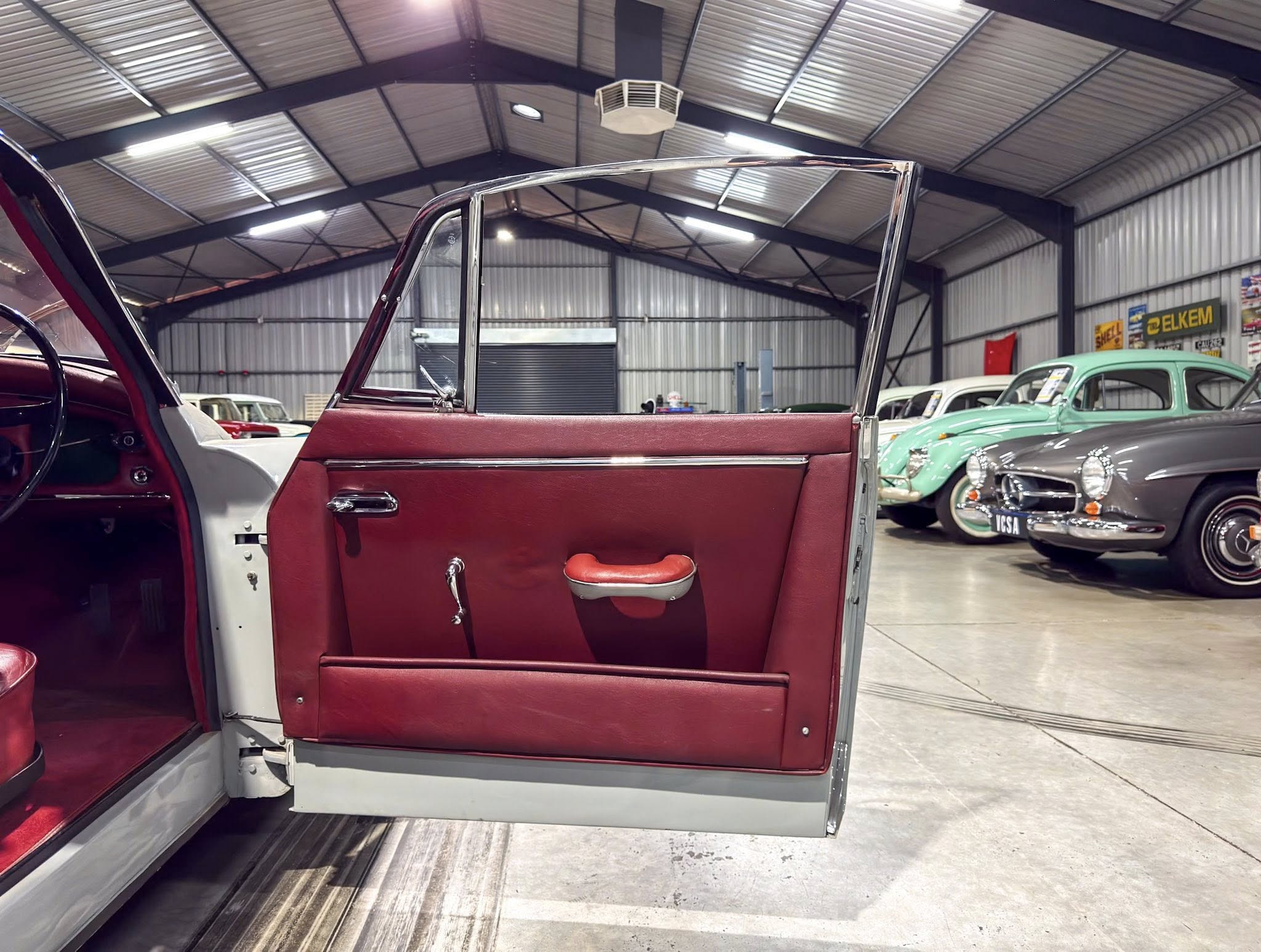 Open red vintage car door revealing its interior, with other classic cars on display in a spacious metal showroom.