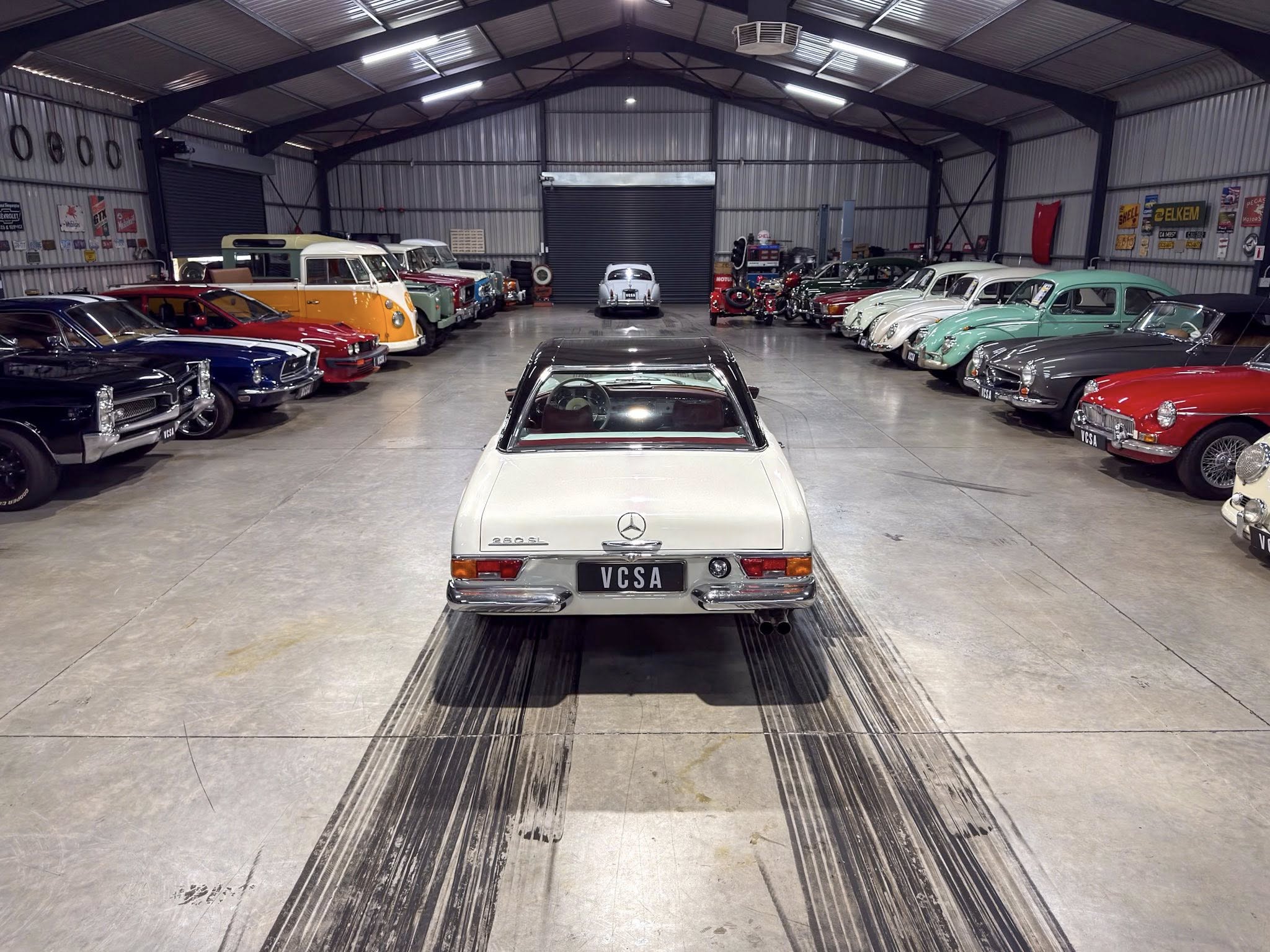 Wide view of a vintage car showroom/garage with rows of classic cars on both sides and a white Mercedes in the foreground, VC SA plate visible.