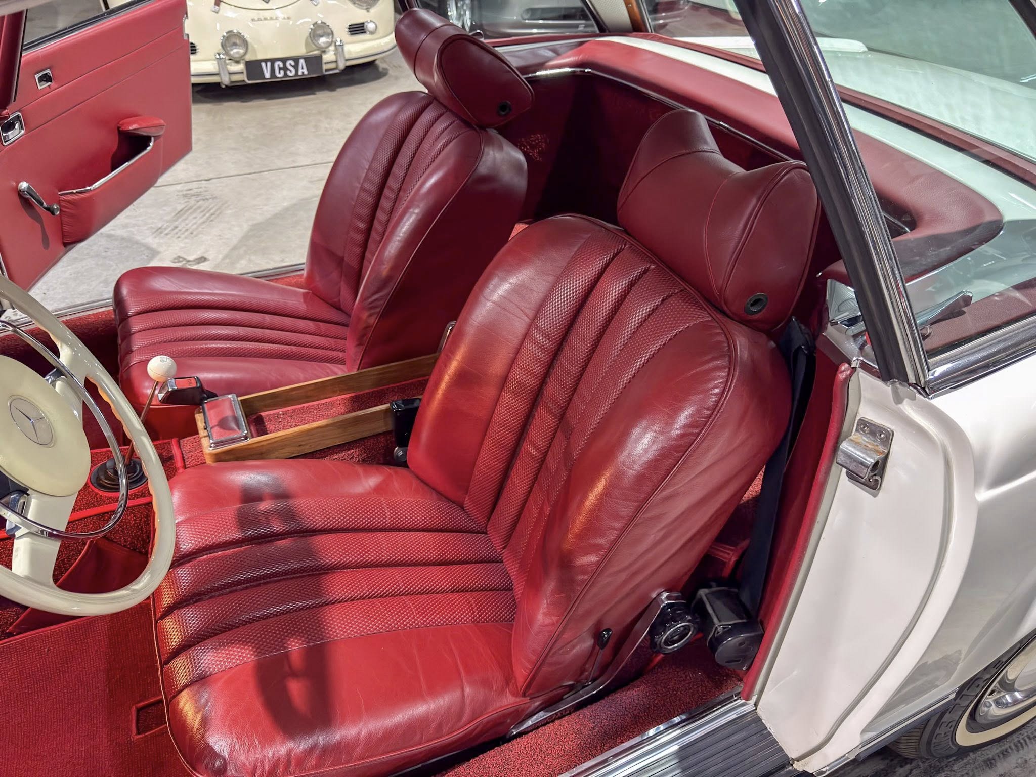 Red leather car interior with front seats, steering wheel, and wood center console; door open in a vintage car.