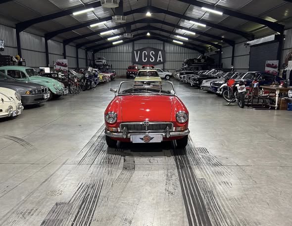 Red vintage convertible centered in a large indoor showroom filled with classic cars on both sides; VCSA sign on the back wall.