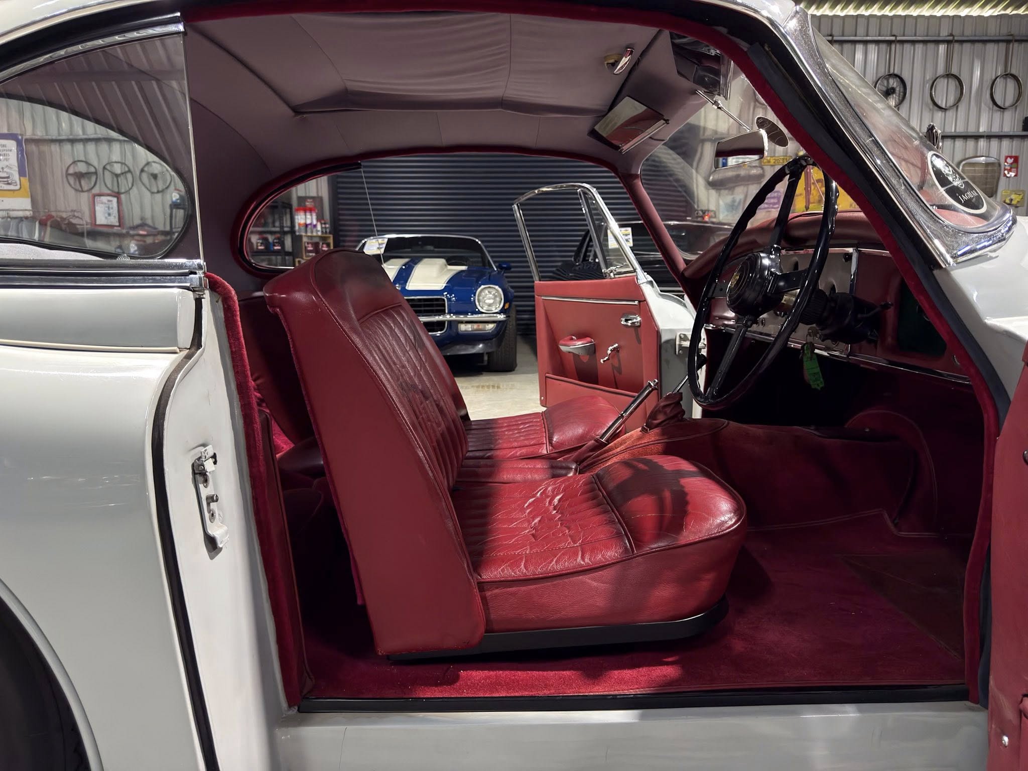 Inside a vintage car with red leather seats and door panels, open driver's door, showing a classic steering wheel in a dim garage with another blue car in the background.