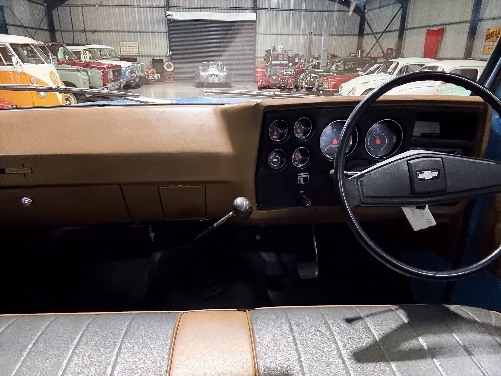 View from the driver’s seat of a vintage car, showing a brown dashboard, circular gauges, and a black steering wheel in a classic car showroom filled with colorful retro vehicles behind a windshield.