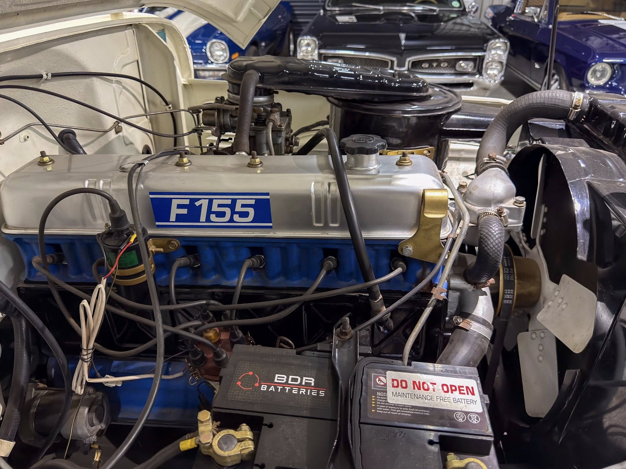 Engine bay of a classic car with a silver valve cover and blue block labeled F155, surrounded by hoses and wiring and a maintenance-free battery nearby.