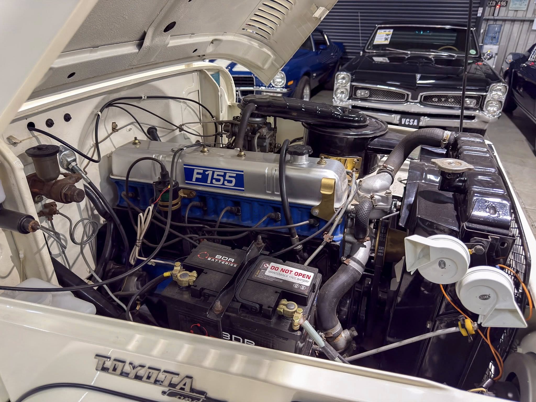 Open hood of a classic Toyota car showing its engine bay, with a blue engine block labeled F155 and a visible TOYOTA badge, in a showroom with other vintage cars in the background.