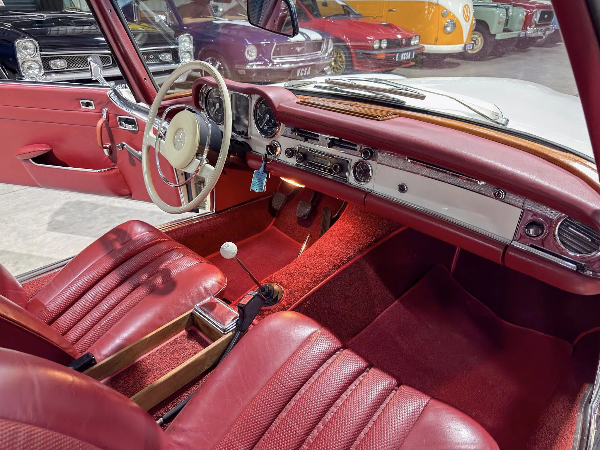 Interior of a red vintage car with a cream steering wheel, wood dashboard trim, and red leather seats; other classic cars visible through the windshield.