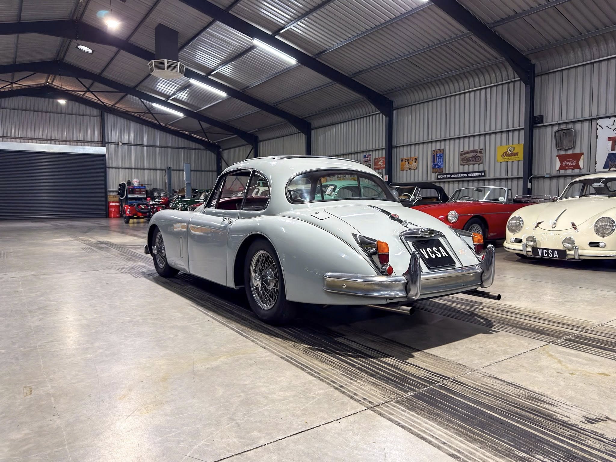 Indoor showroom/garage filled with vintage cars; the foreground features a silver classic coupe with rounded rear, others in red and cream along the wall.