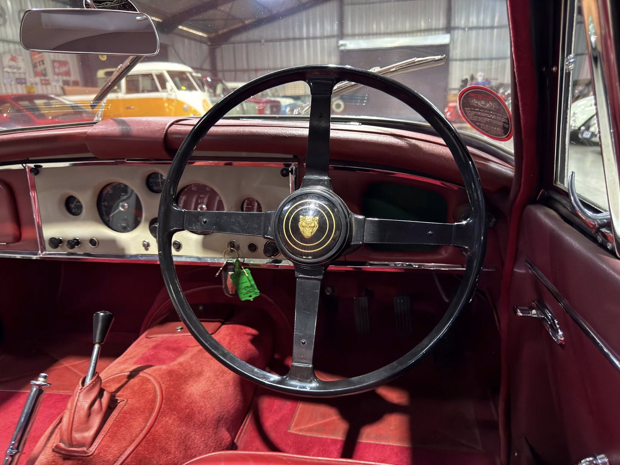 Vintage car interior: red leather dashboard and large black steering wheel with gold emblem, showroom of classic cars visible outside windshield.