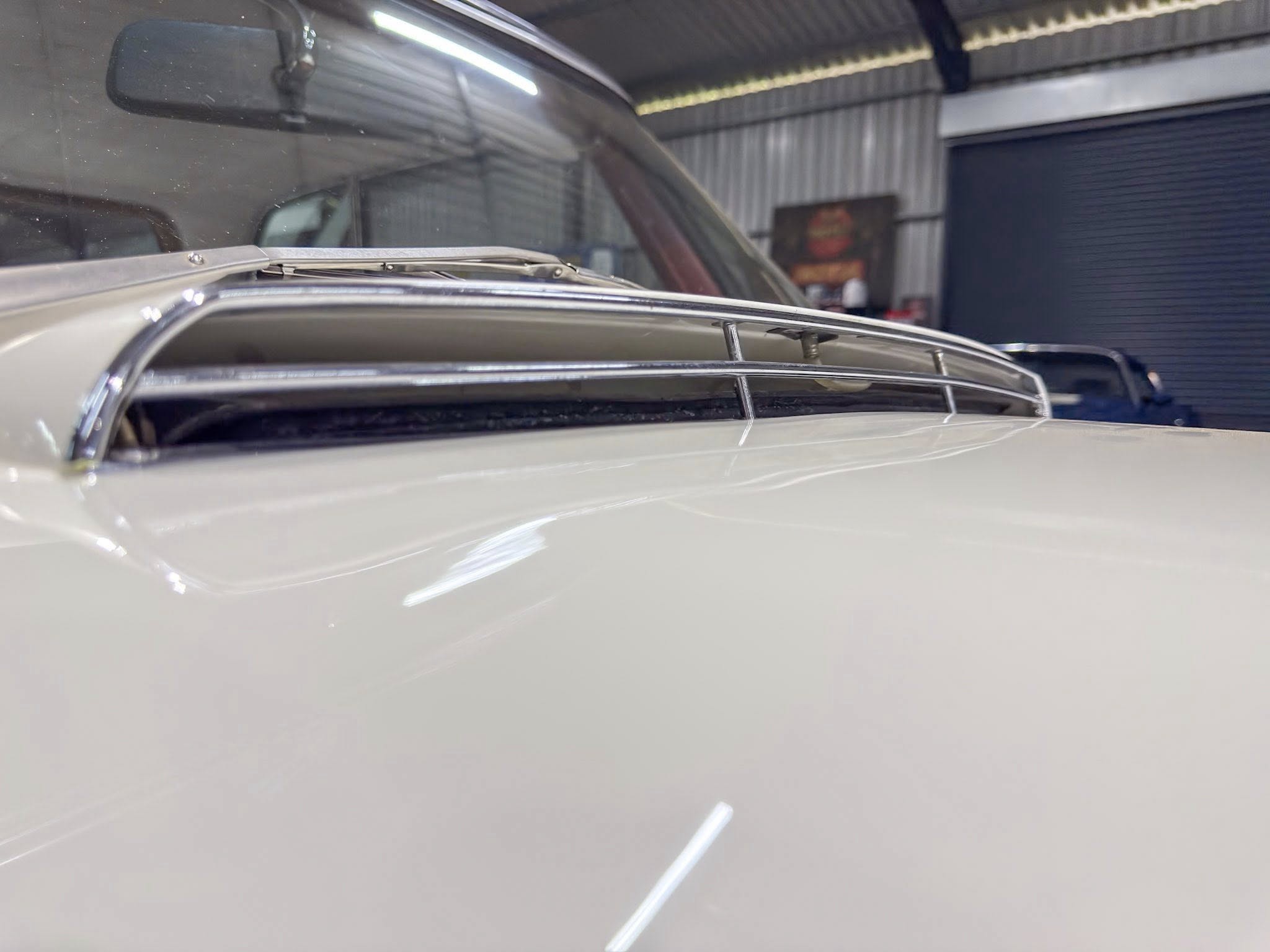 Close-up of a vintage car's chrome roof rack and windshield area inside a dim garage, with soft reflections on the glossy paint