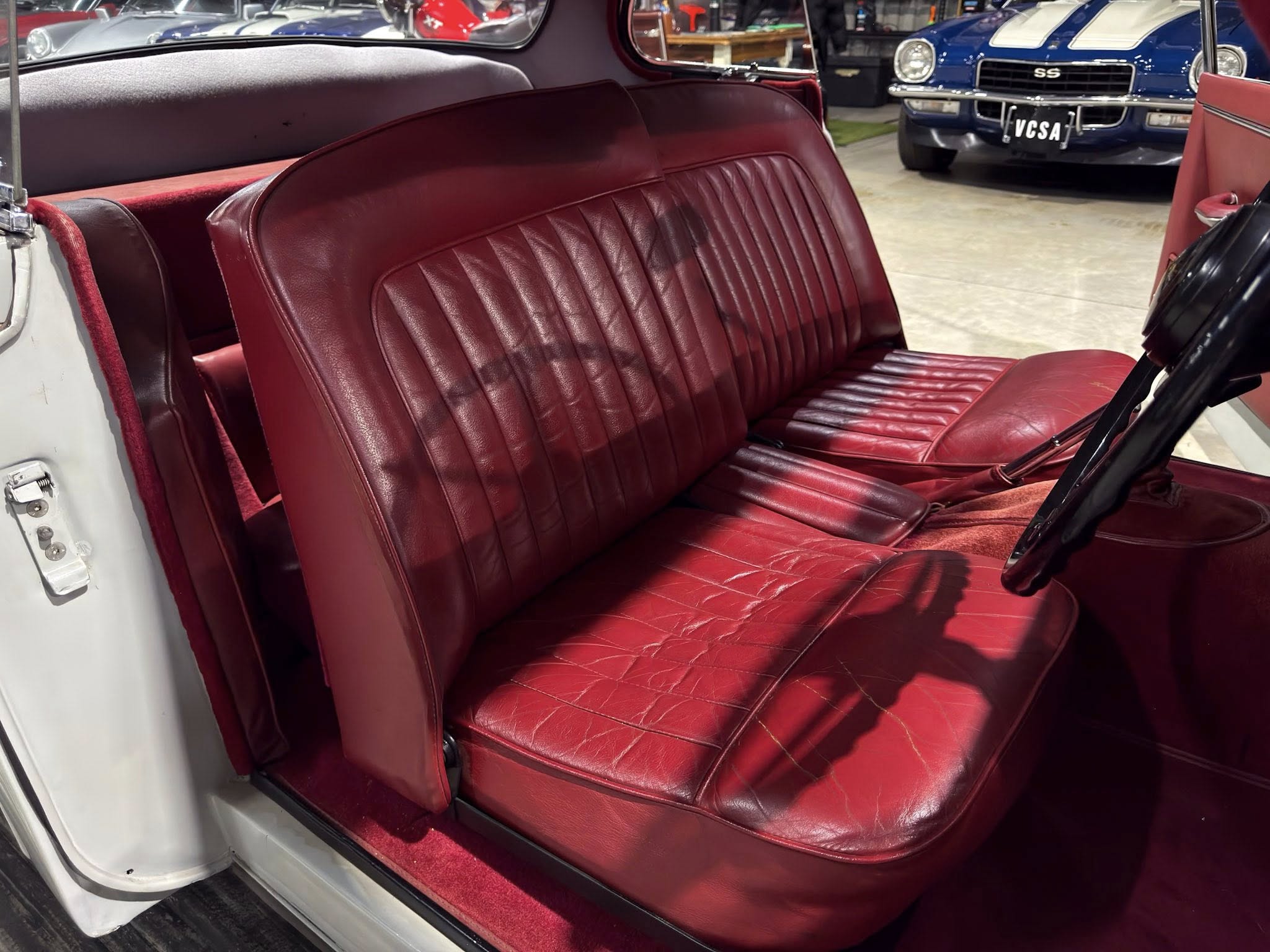 Red leather front and bench seats inside a vintage car interior, with red carpet and a visible door frame on the left and a classic blue car in the background.