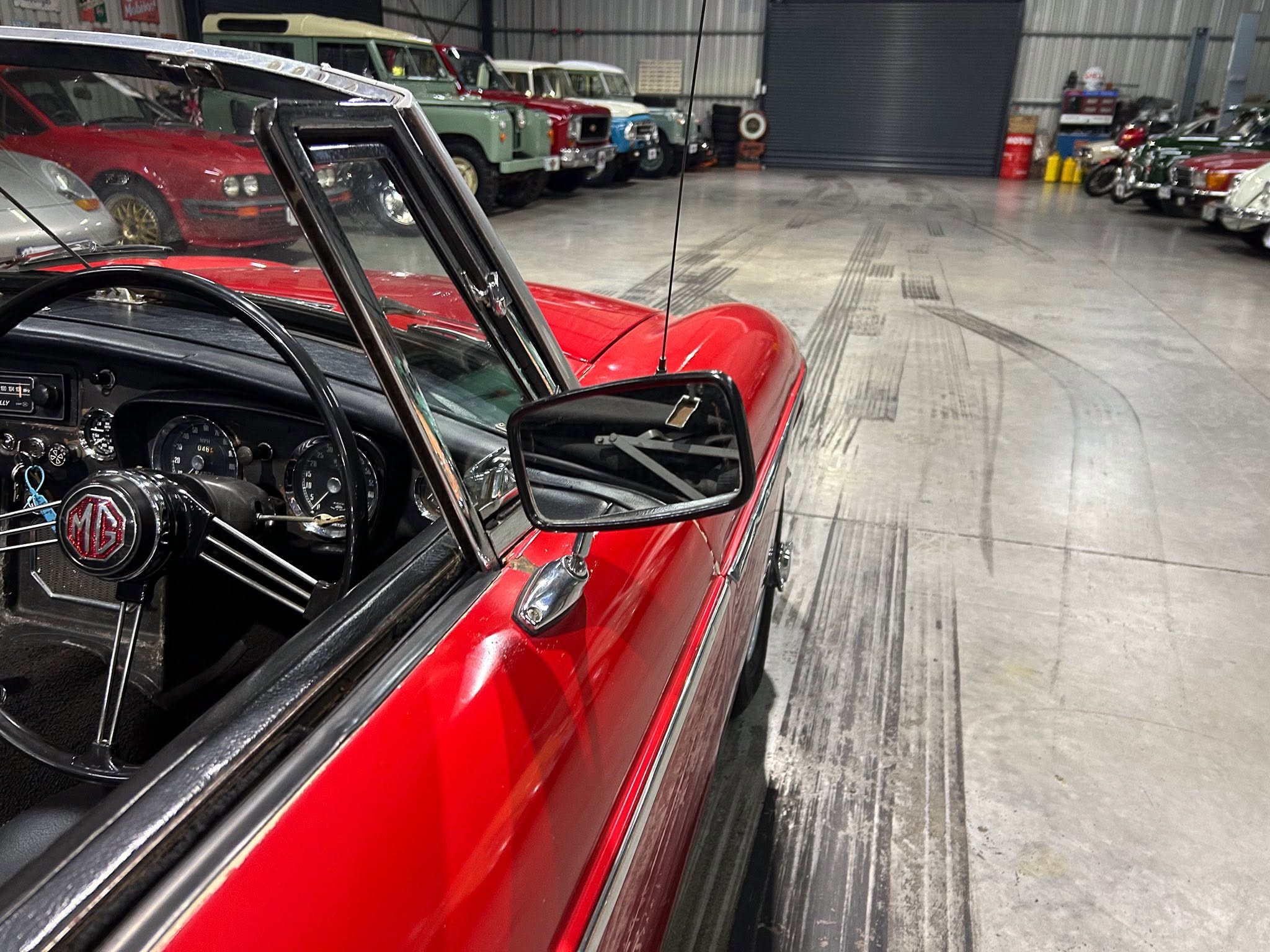 Red vintage MG car interior with steering wheel and dashboard, door open, in a showroom full of classic cars in the background.